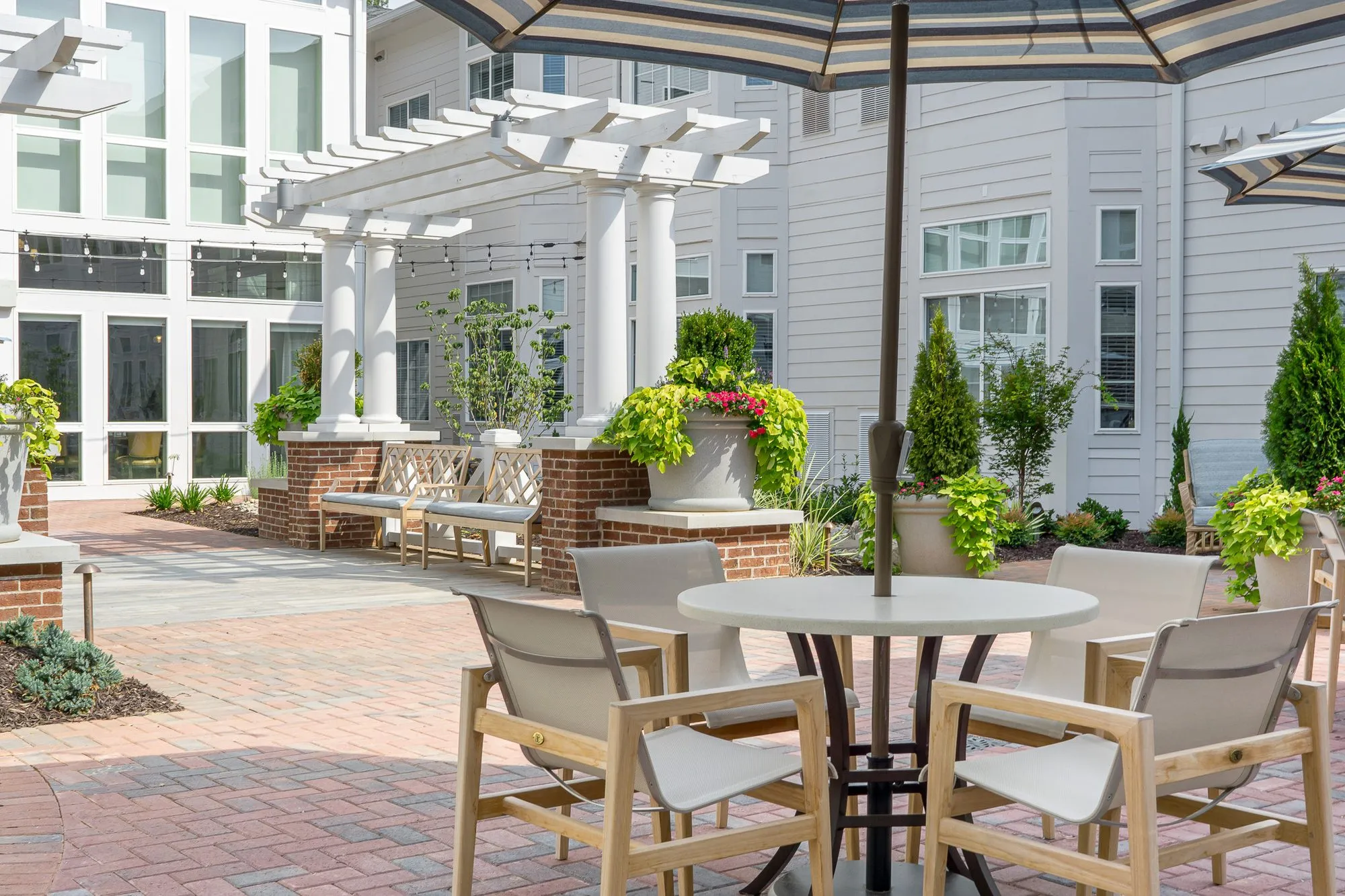 Senior living courtyard with teak dining chairs at a round table under a striped umbrella, a pergola and brick planter beyond