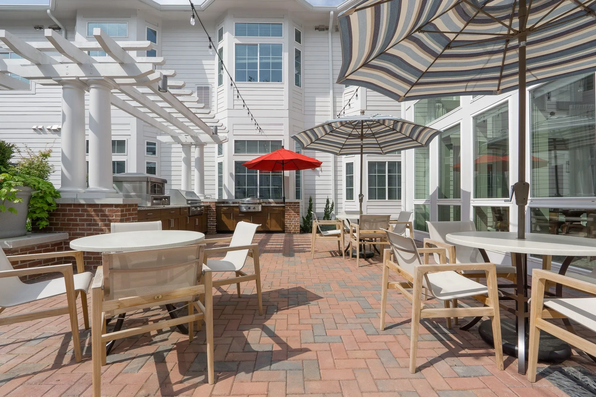 Senior living courtyard with sling chairs at round tables beside the glass-walled wing and pergola-covered outdoor kitchen