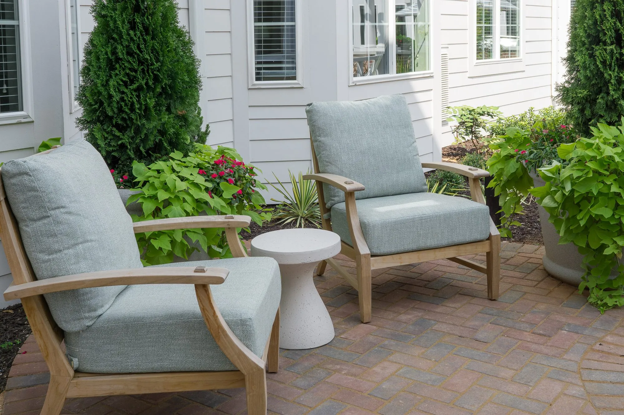 Senior living outdoor seating: two teak armchairs with pale blue cushions and a white side table among planted beds