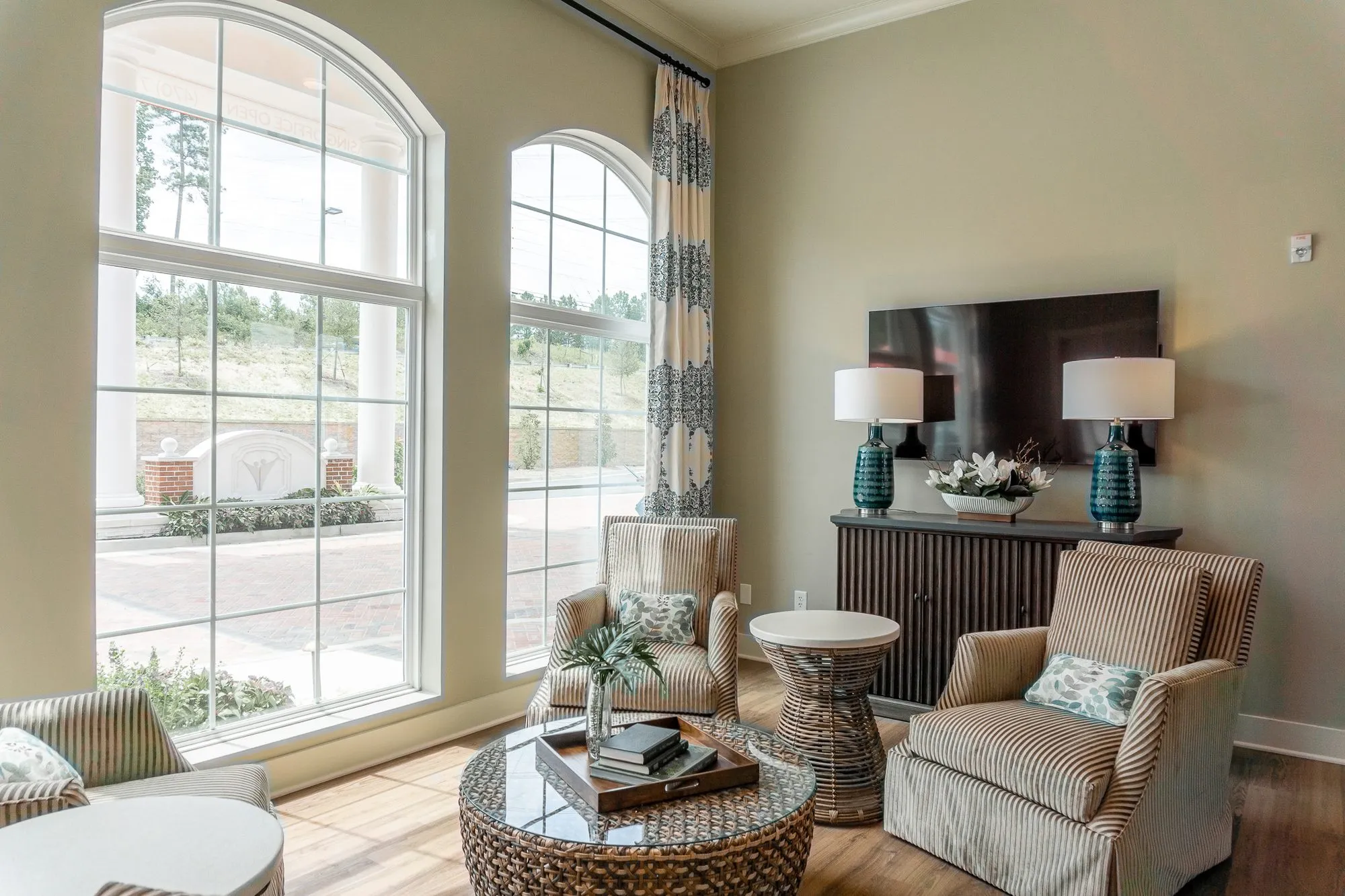 Senior living sitting room with two striped armchairs at a woven coffee table beside tall arched windows, teal lamps