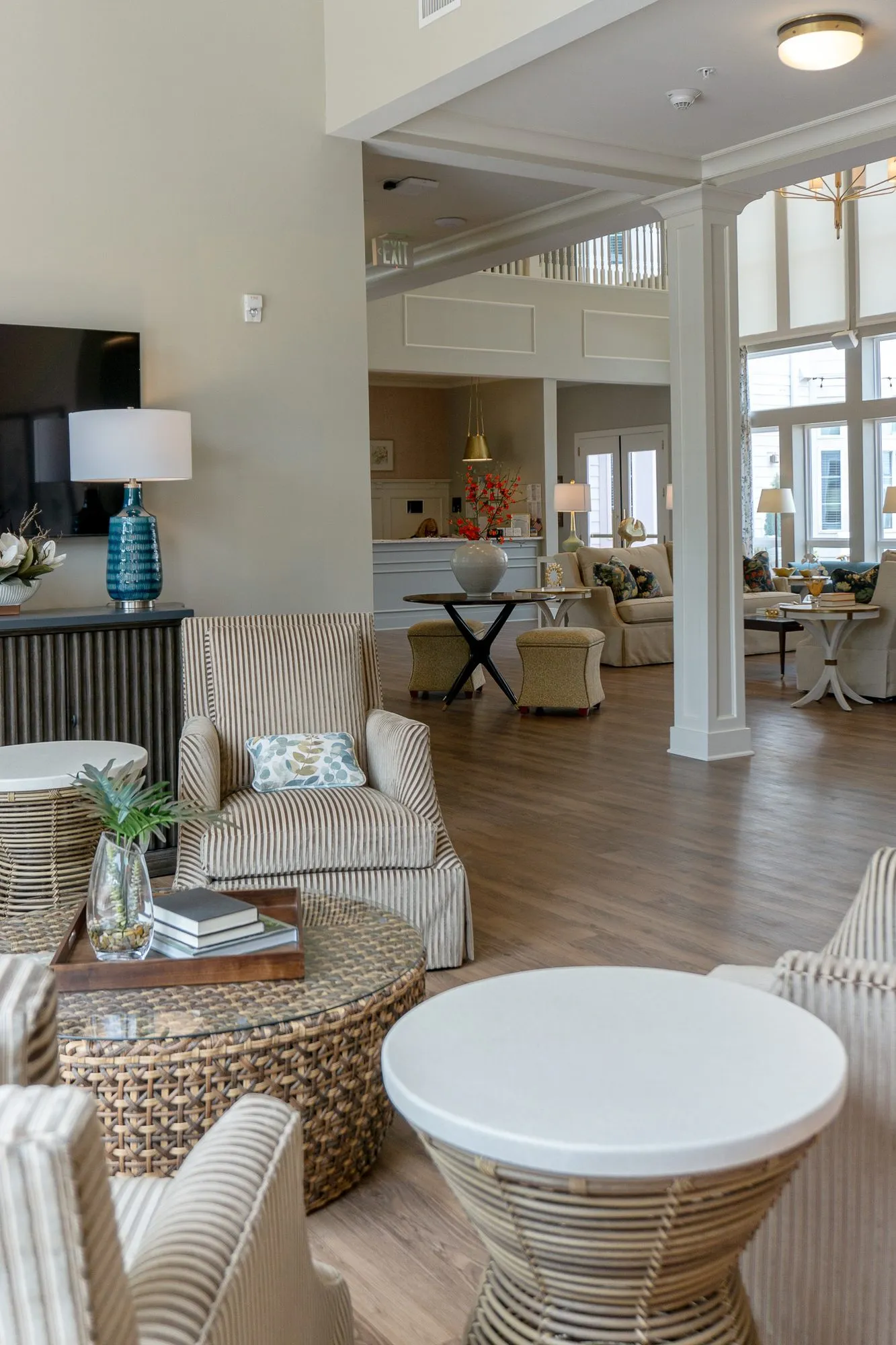 Senior living sitting room seen past striped armchairs and a woven table toward the open great room and kitchen