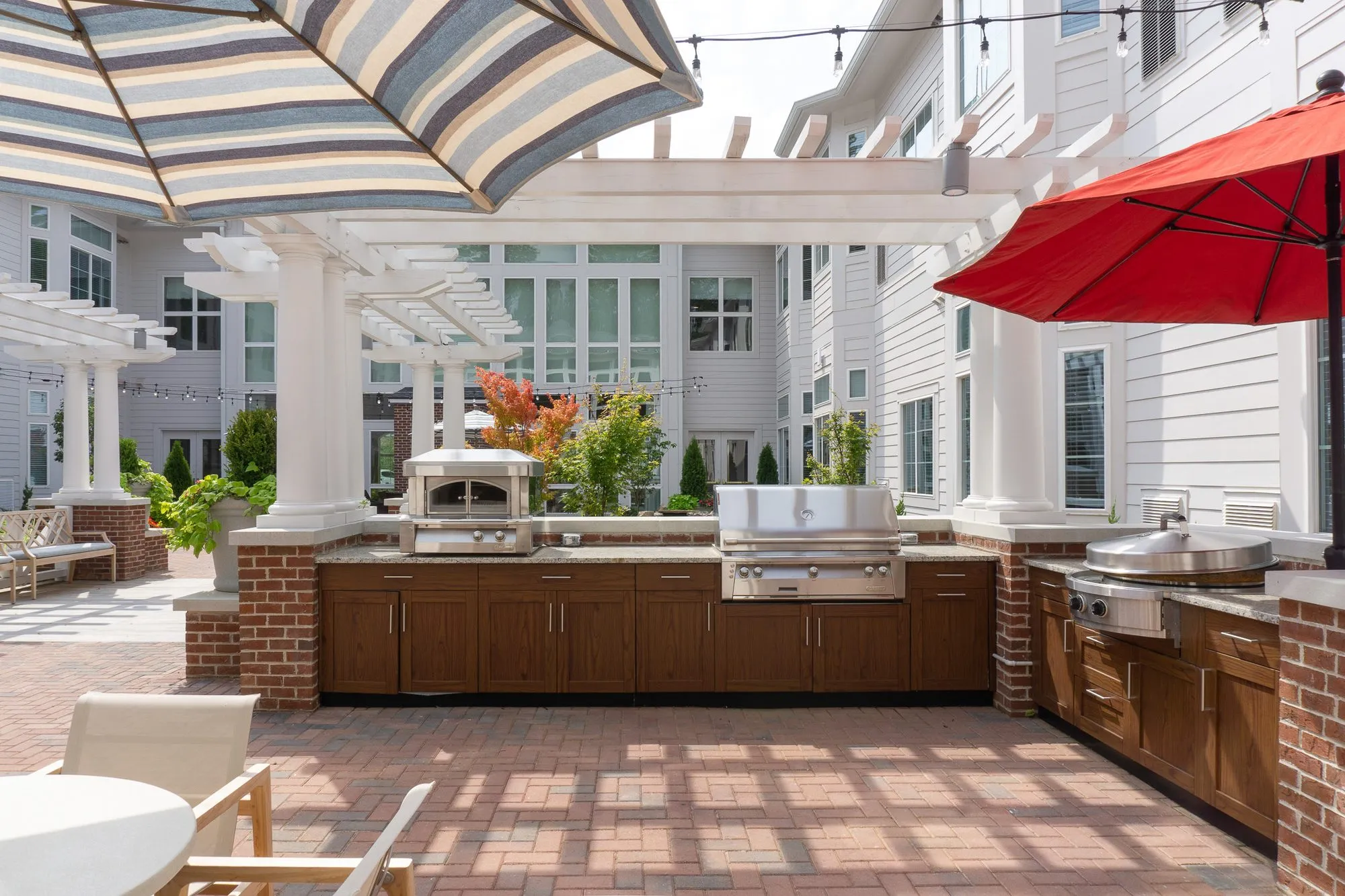 Senior living outdoor kitchen with wood cabinetry, a pizza oven and grills on brick, under striped and red umbrellas