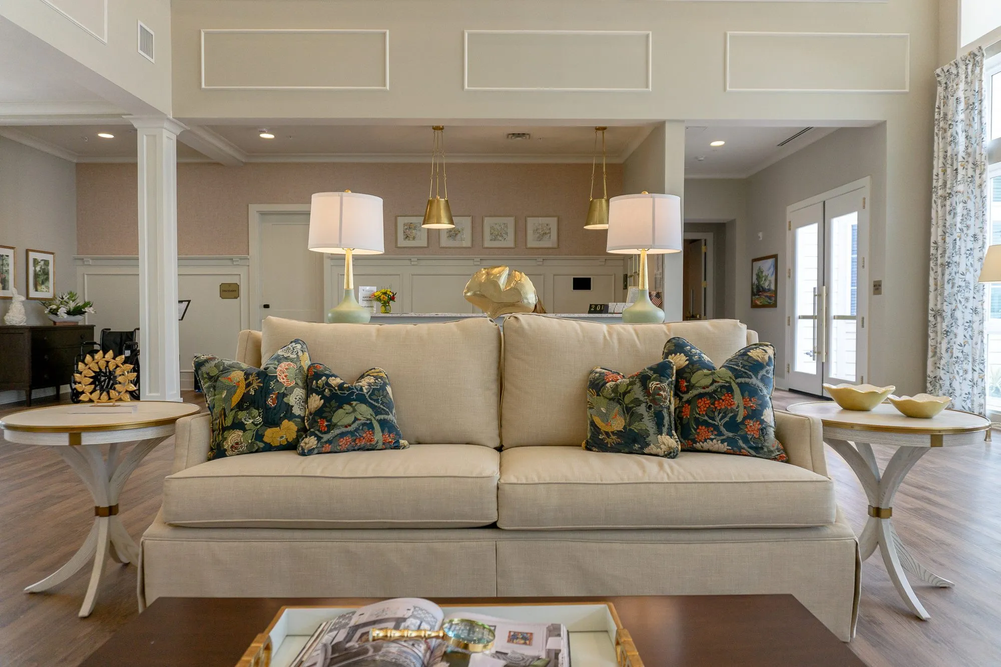Senior living great room with a cream sofa and navy floral pillows between gold-trimmed lamps, a reception counter behind