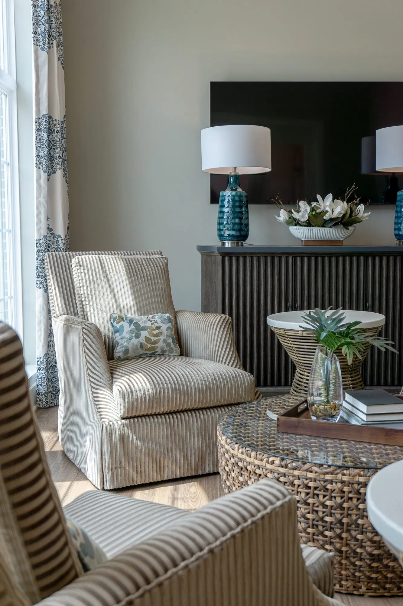 Senior living sitting room with a striped armchair by the window, a woven table and a dark sideboard with teal lamps