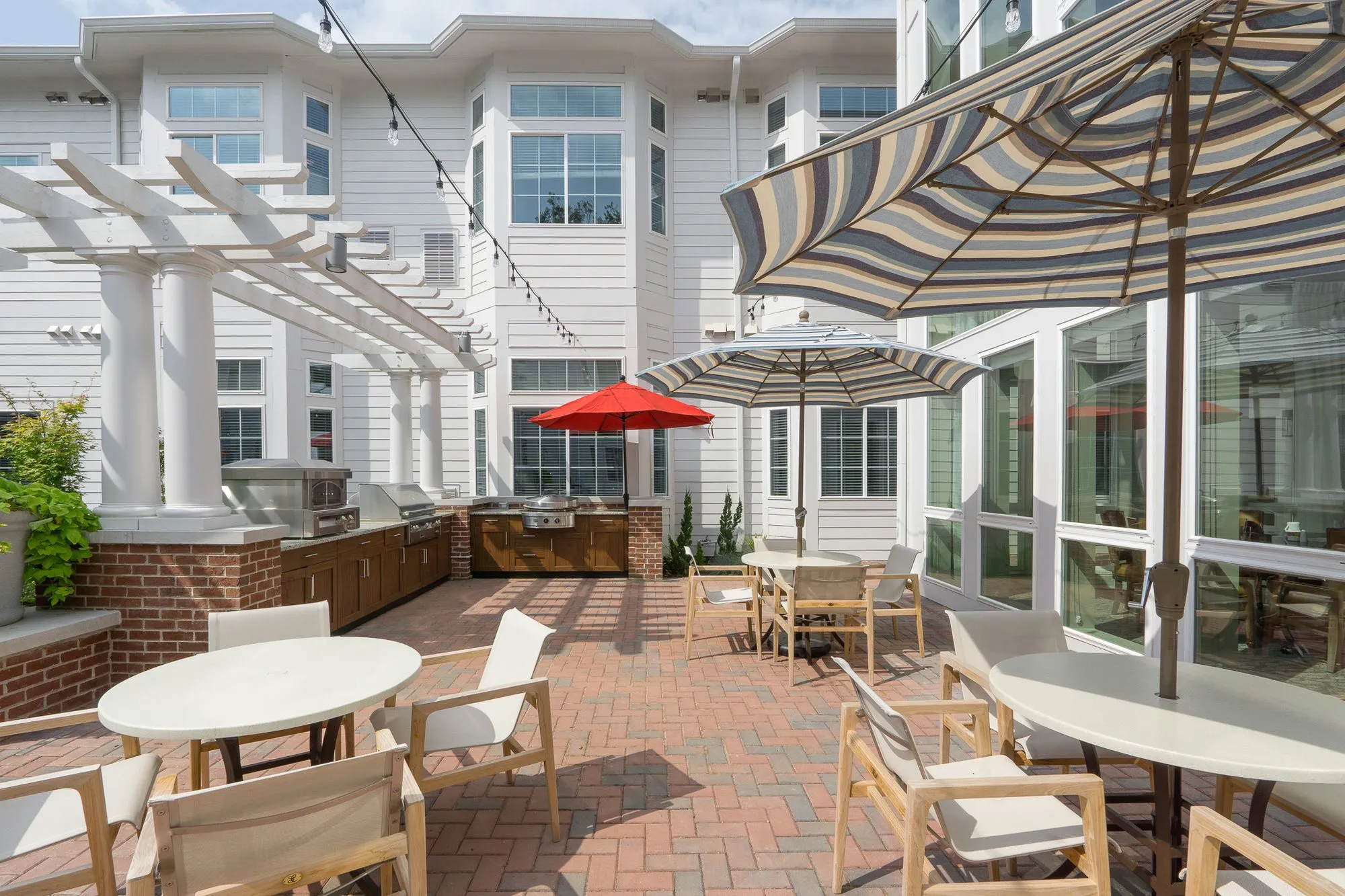 Senior living courtyard with white tables and sling chairs on brick paving leading to the outdoor kitchen and red umbrella