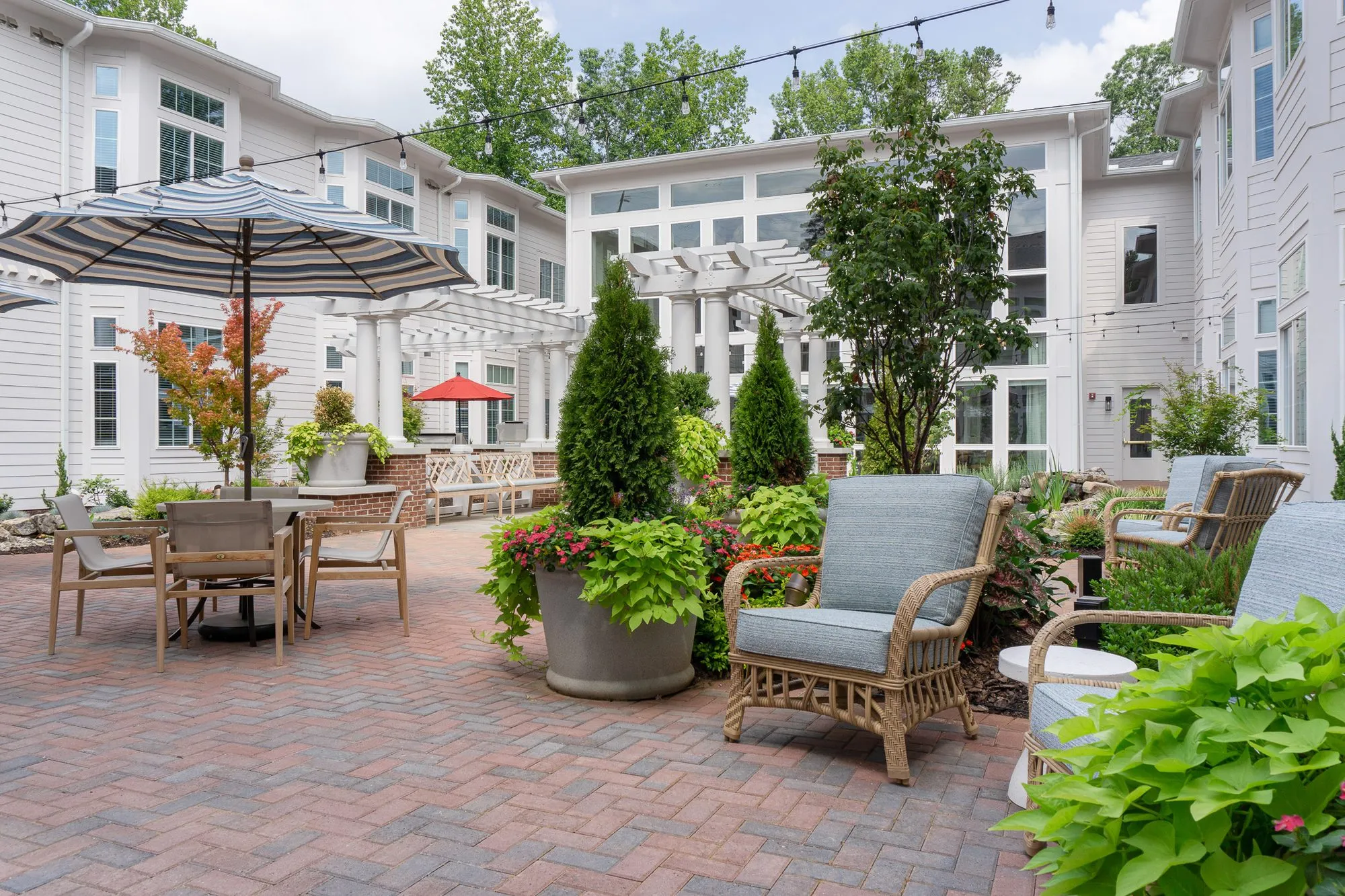 Senior living courtyard with a blue-cushioned rattan armchair beside a planter of annuals, cypress and a pergola behind