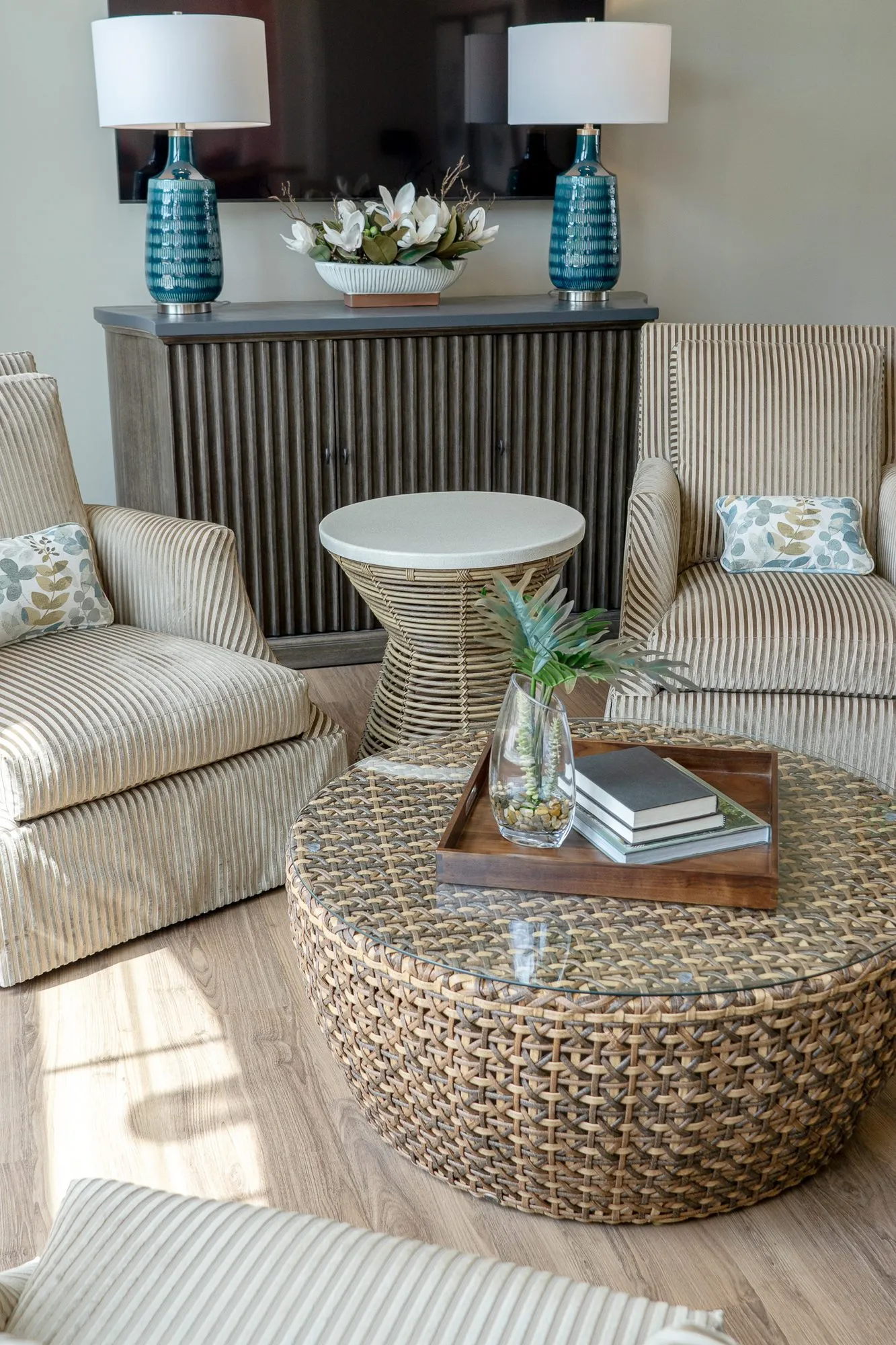 Senior living sitting room from above, a woven drum table with a tray of books before two striped armchairs and teal lamps