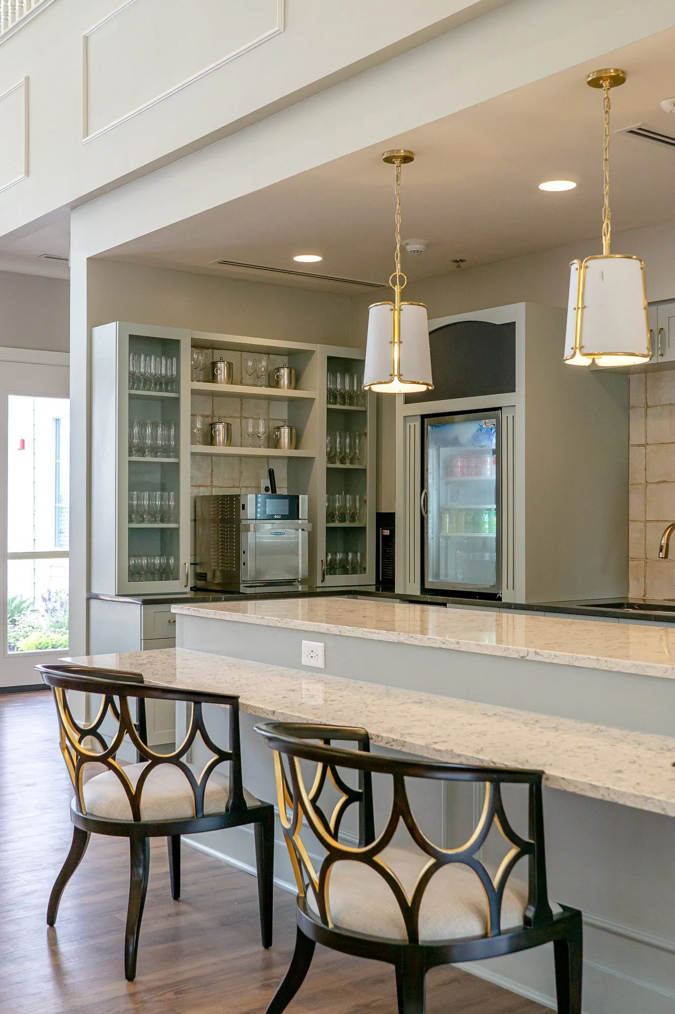 Senior living kitchen counter with black and gold fretwork back chairs under linen pendants, glass-front cabinets beyond
