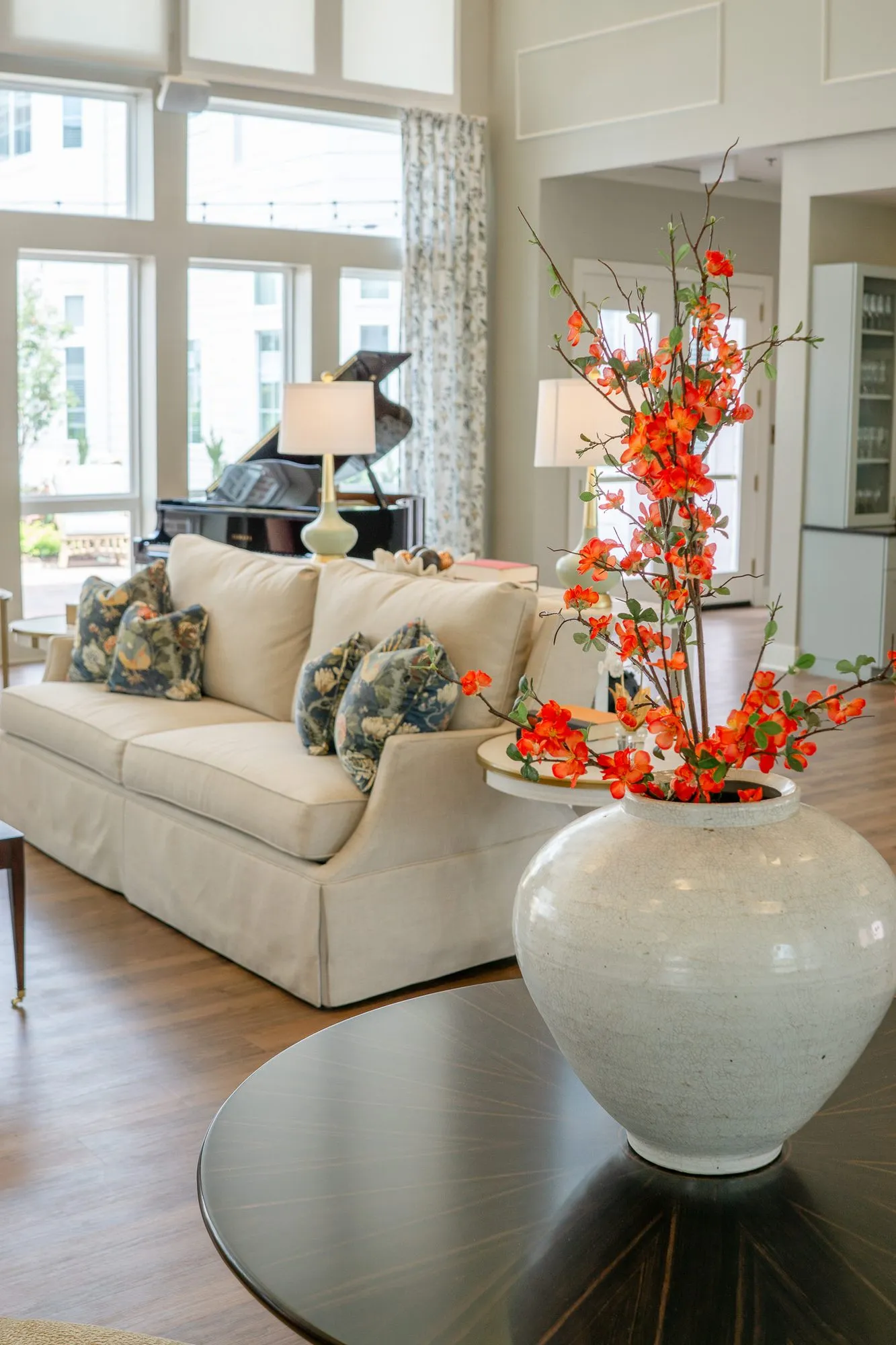 Senior living great room with a vase of orange blossom on a dark round table, a cream sofa and grand piano beyond
