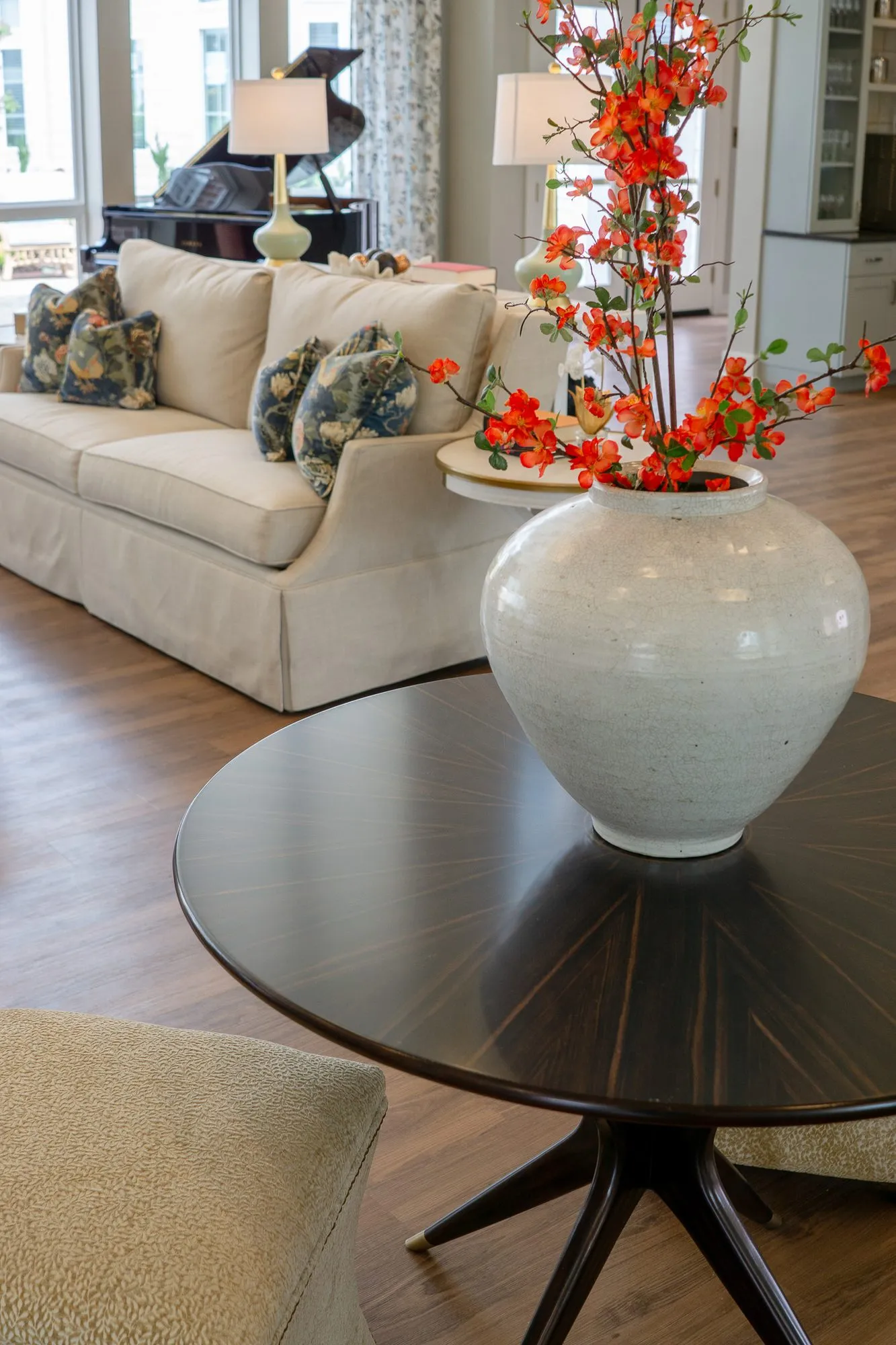 Senior living interior detail: white vase of orange blossoms centred on a dark round table, the cream sofa behind