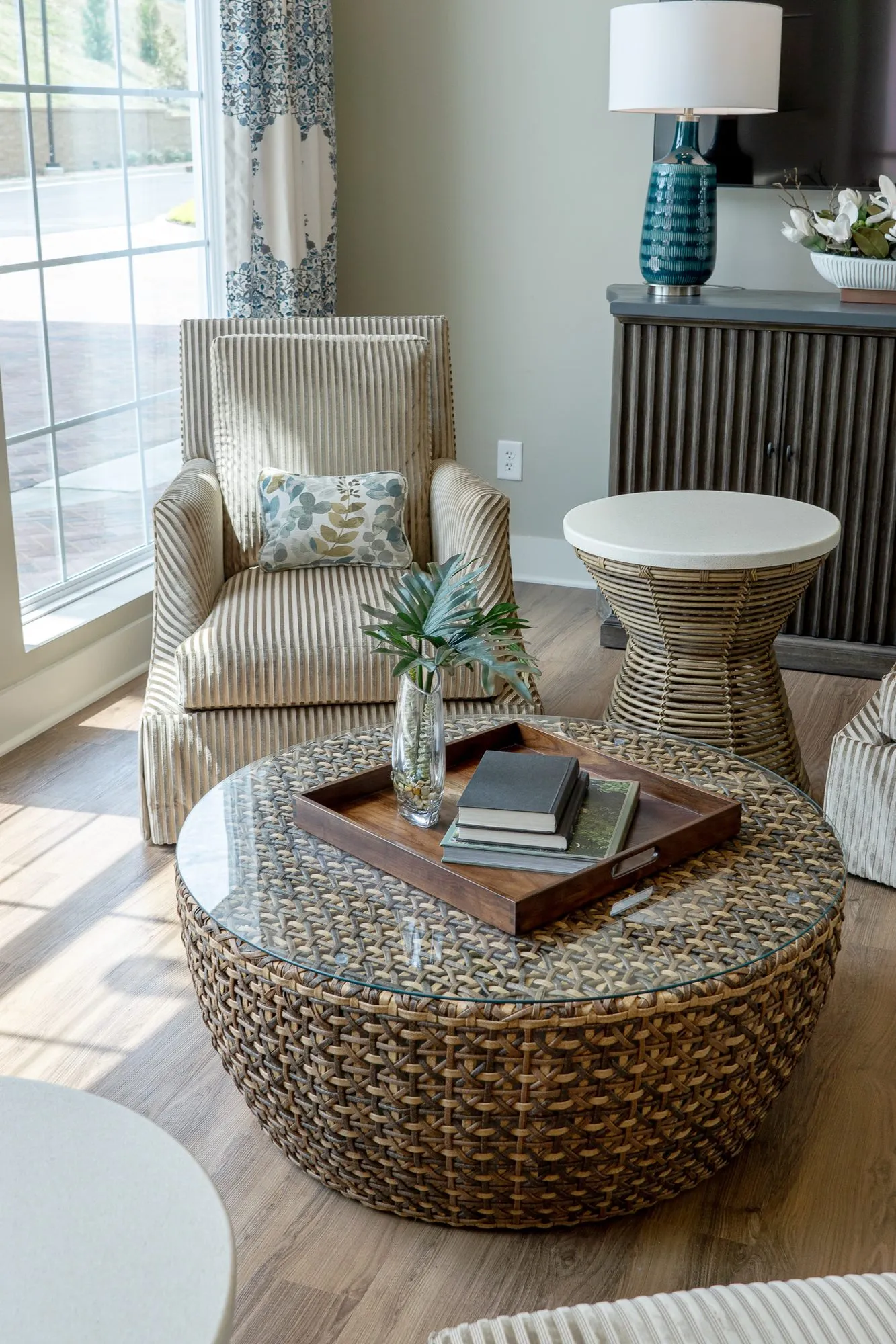 Senior living sitting room with a woven drum coffee table and books before a striped armchair, a teal lamp behind