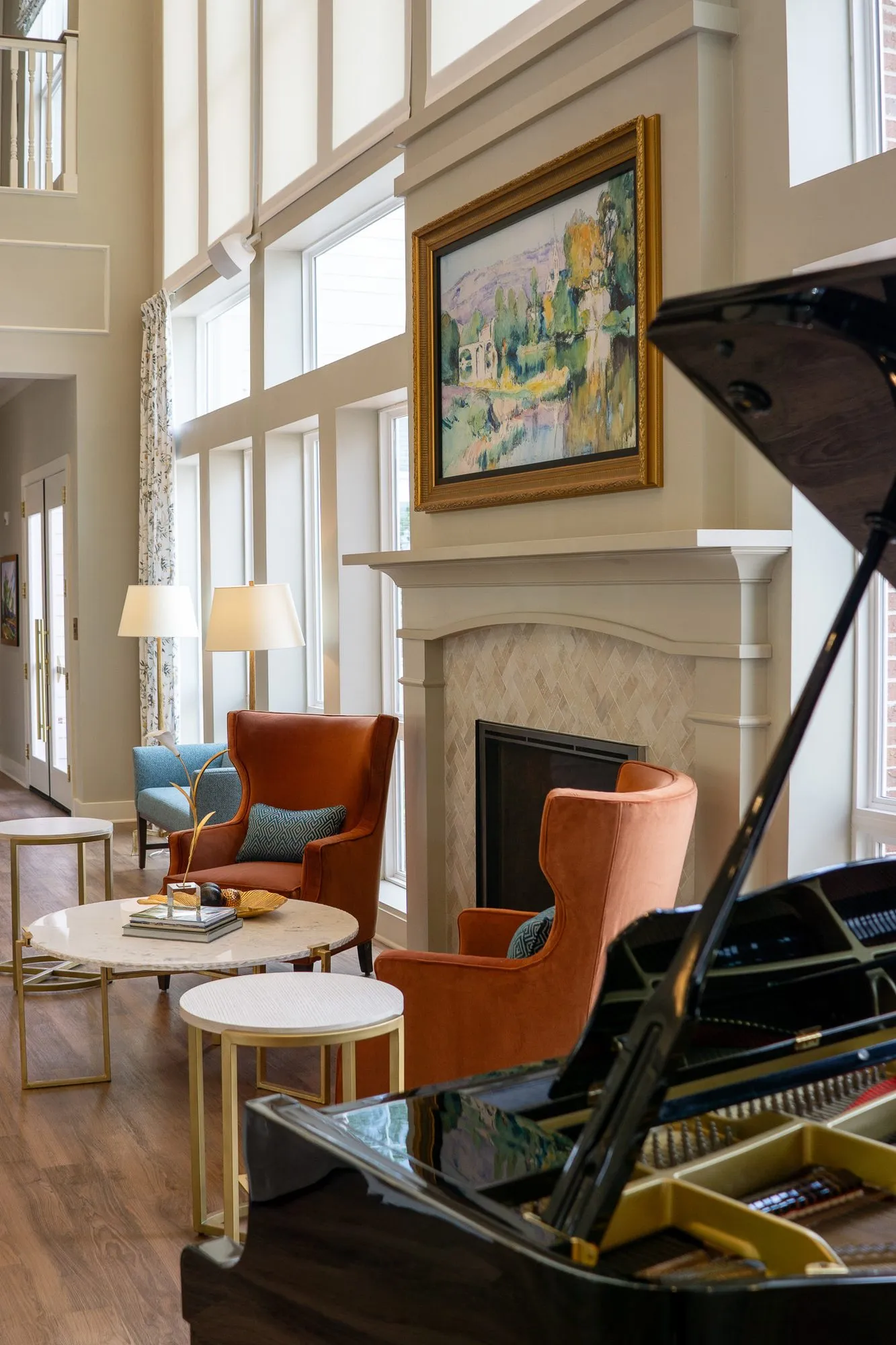 Senior living interior detail: orange wing chairs at the fireplace below a gilt-framed garden painting, a piano lid in front