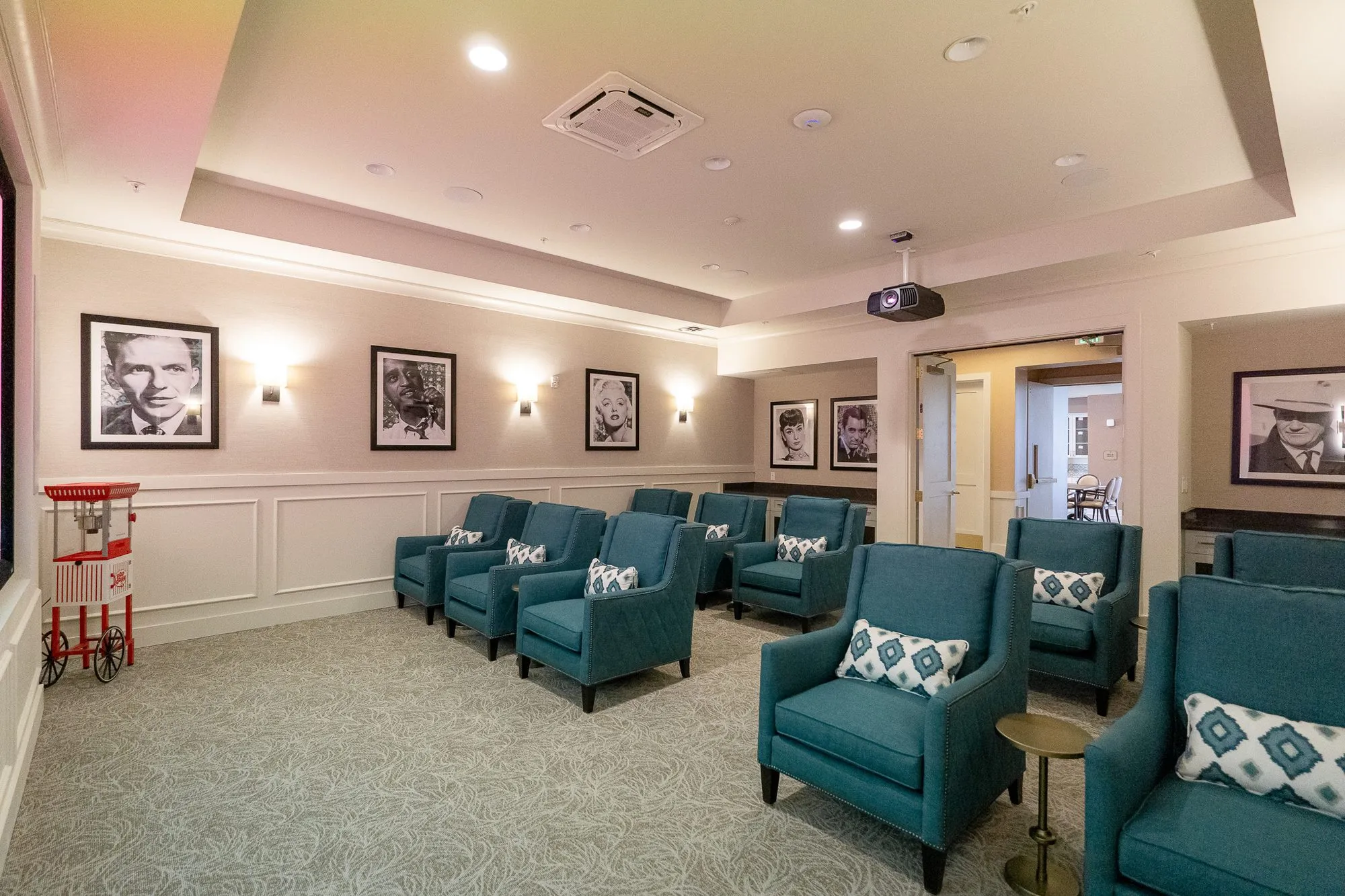Senior living theater with rows of teal armchairs on grey carpet, framed black and white portraits and a red popcorn cart