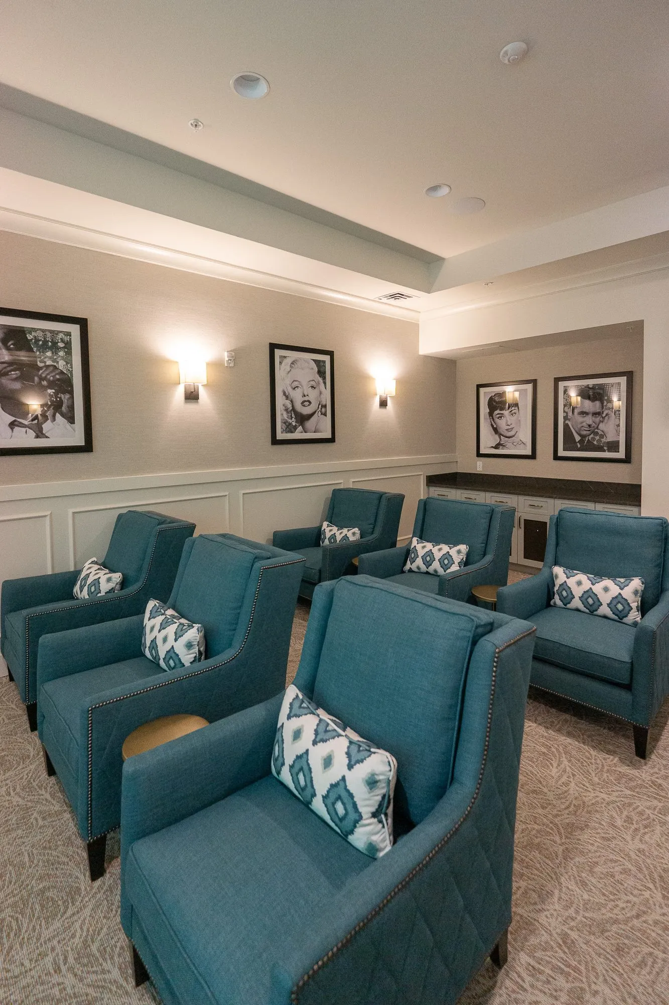 Senior living theater with teal armchairs and diamond print pillows in rows below black and white portraits and sconces