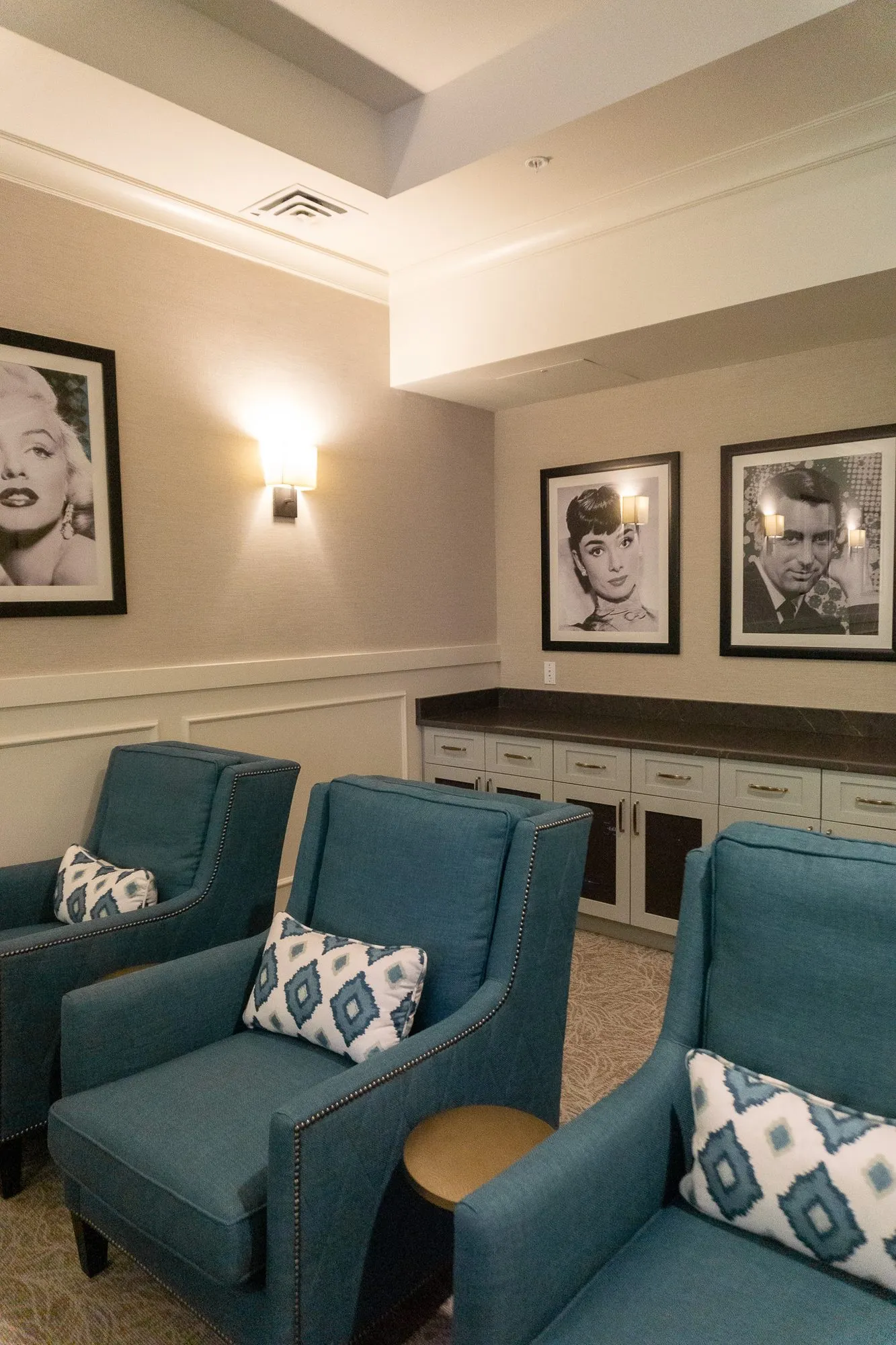 Senior living theater side wall of black and white portraits above teal armchairs and a low cabinet