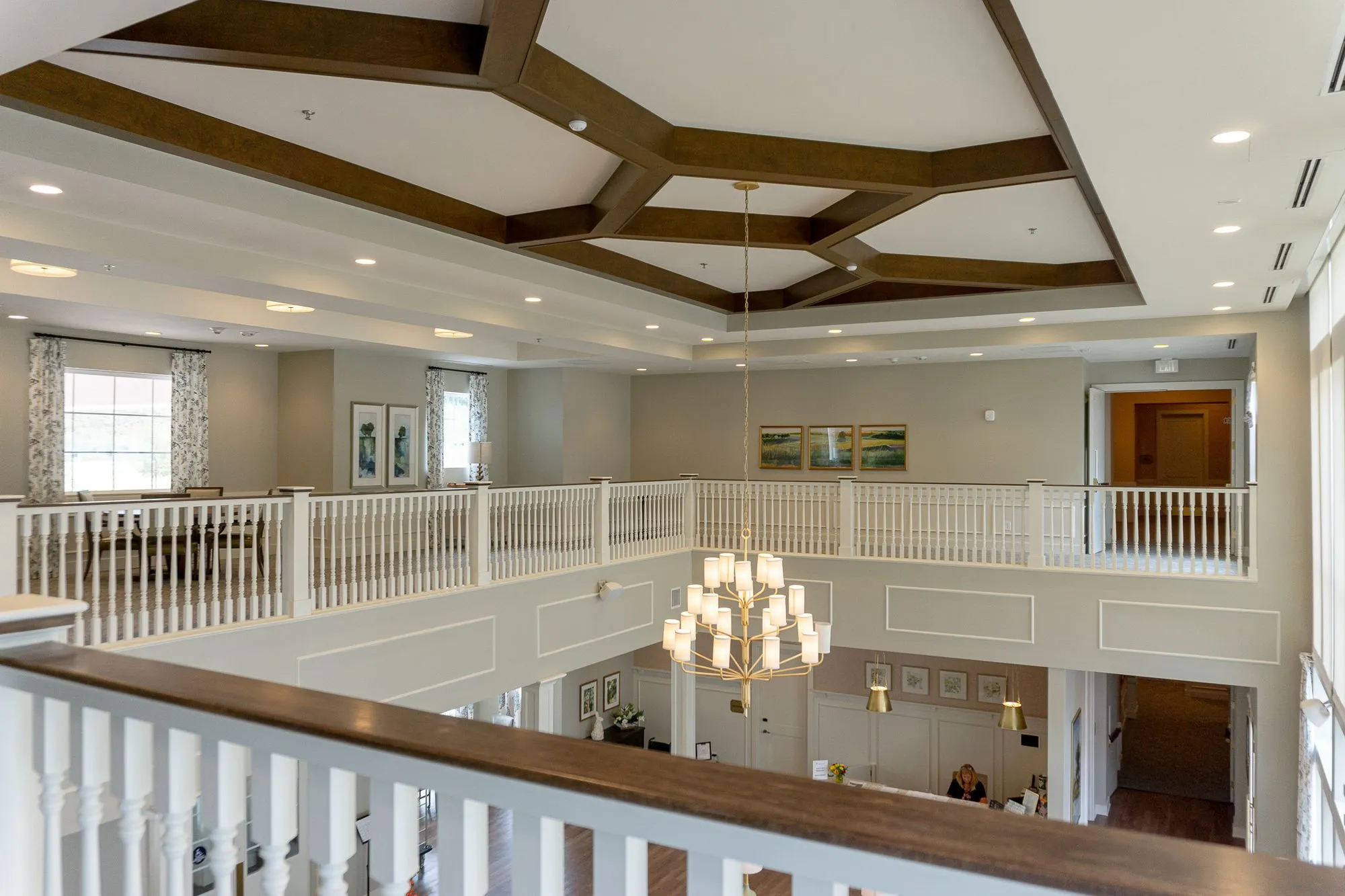 Senior living great room seen from the second-floor balcony, a tiered chandelier under a dark beamed ceiling