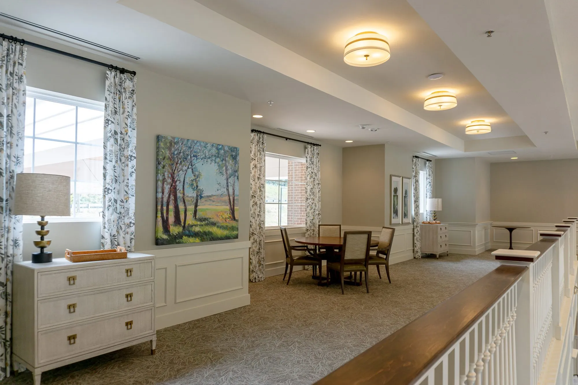 Senior living second-floor landing with a white dresser, a landscape painting and a table for four by the balcony rail