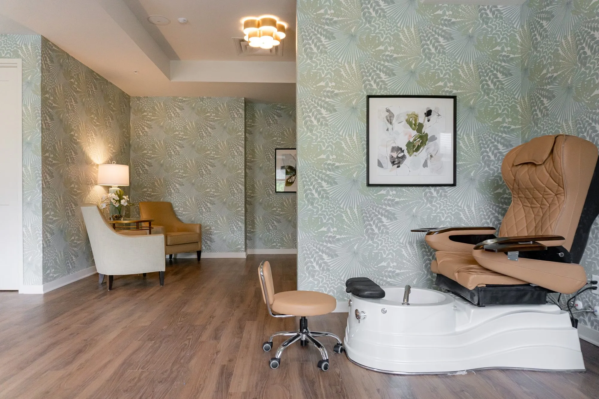 Senior living salon with a tan leather pedicure chair and white basin before palm frond wallpaper, armchairs in the corner