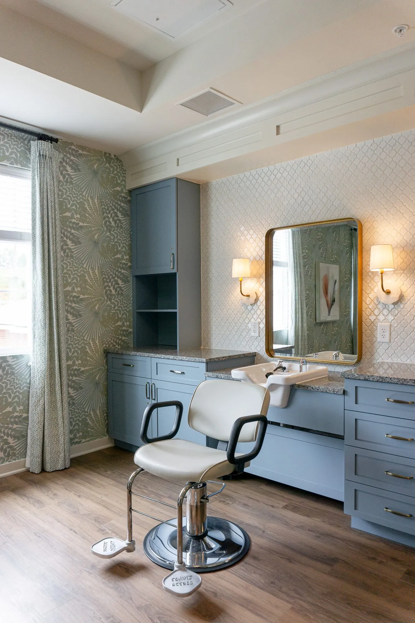 Senior living salon with a white styling chair at a slate blue vanity, a brass mirror and sconces on white arabesque tile