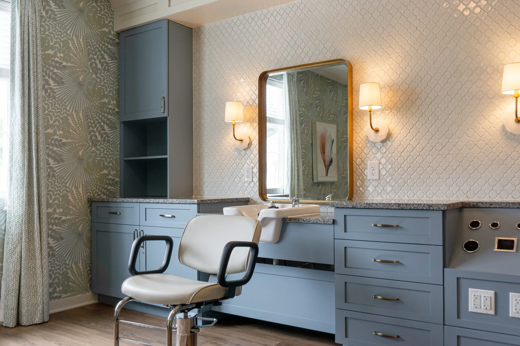 Senior living salon with a white styling chair at a slate blue vanity, a brass-framed mirror between linen sconces