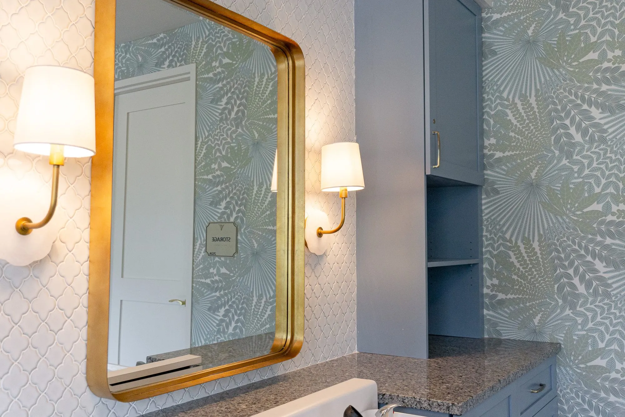 Senior living salon detail: brass-framed mirror and sconces on arabesque tile beside a slate blue cabinet and palm wallpaper