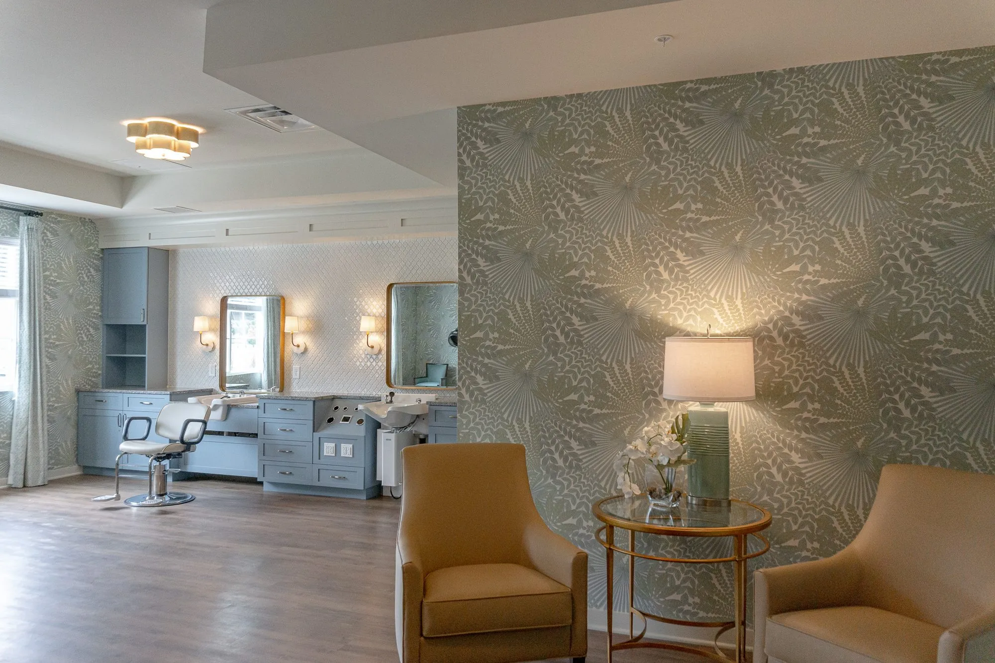 Senior living salon seen past two camel armchairs and a lamp table against palm frond wallpaper, styling stations beyond