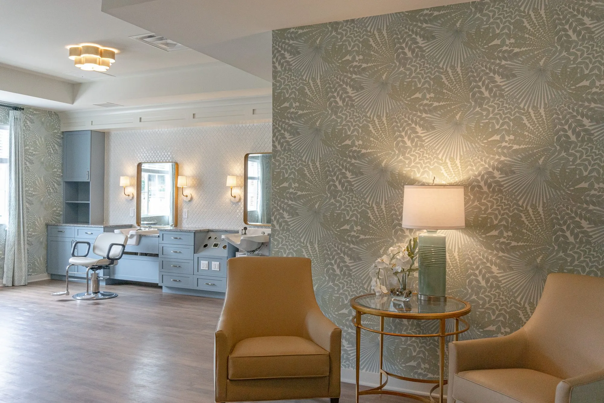 Senior living salon with two camel armchairs flanking a glass side table and lamp on palm frond wallpaper