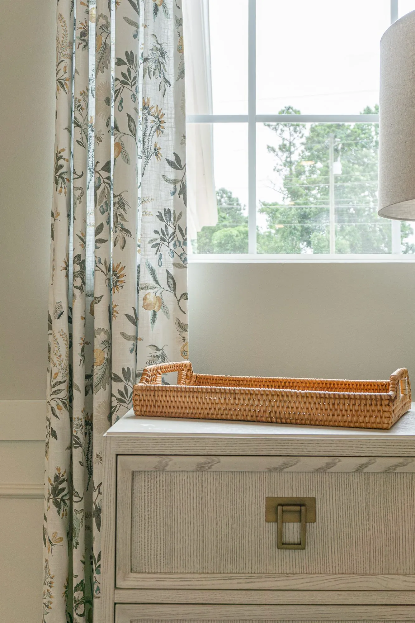Senior living interior detail: whitewashed chest with a woven tray beneath a window with floral drapery