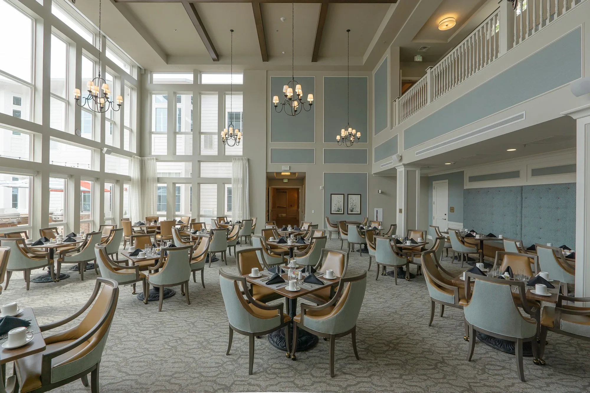 Senior living double-height dining room with set tables, sage and camel chairs, a balcony rail above and tall windows