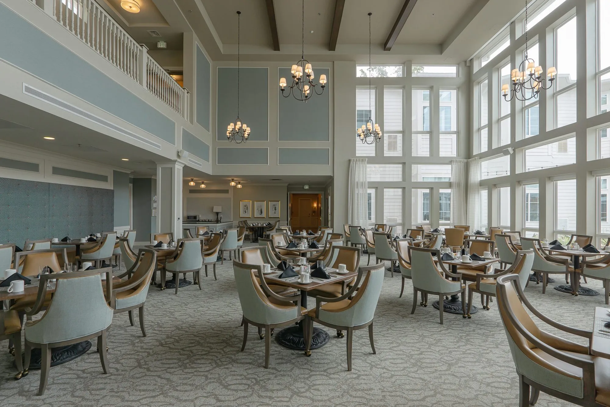 Senior living dining room from beneath the balcony, a panelled sage wall with candle chandeliers above rows of set tables