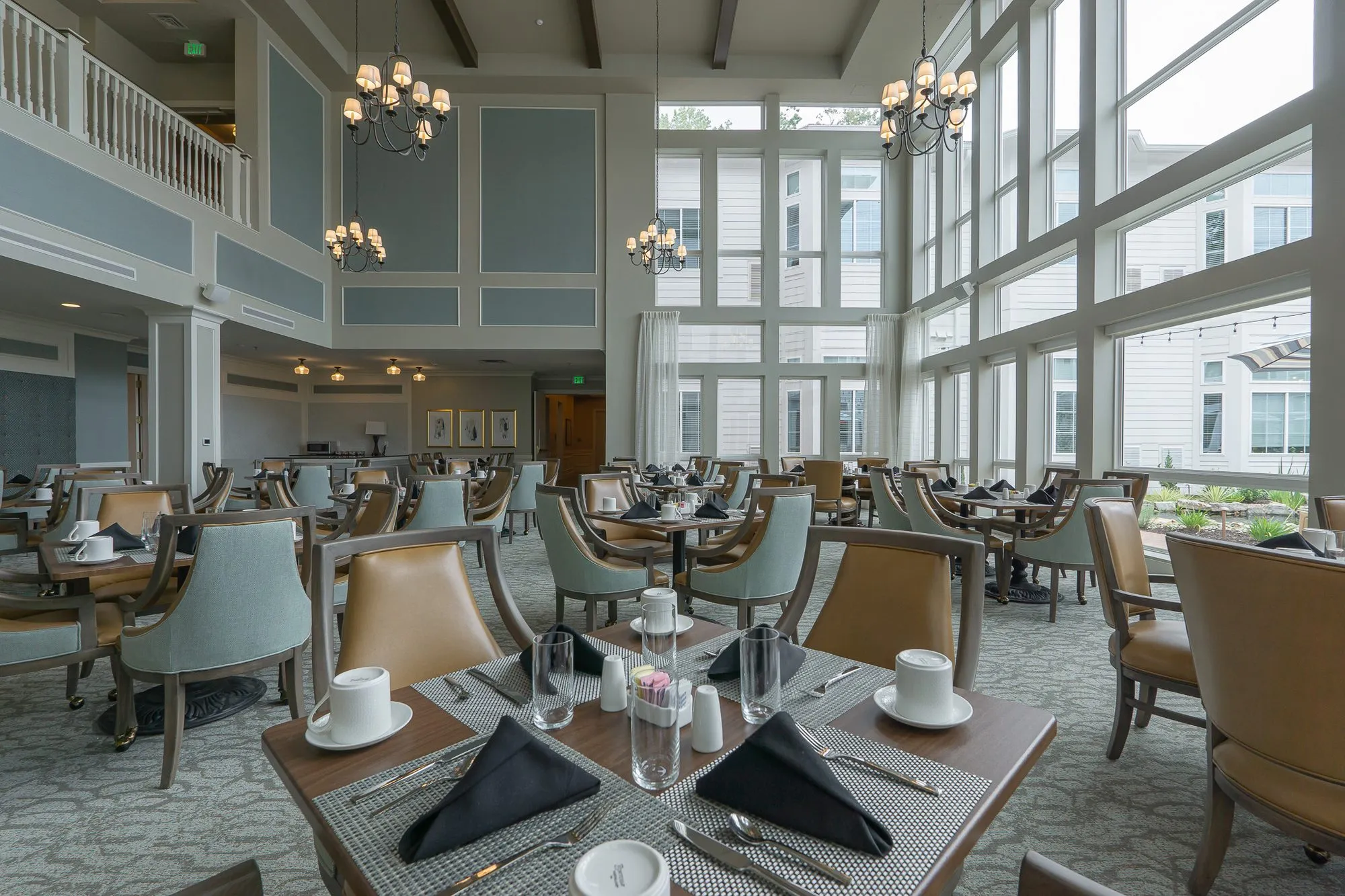 Senior living dining room with a table set with black napkins and white cups, window walls and chandeliers beyond