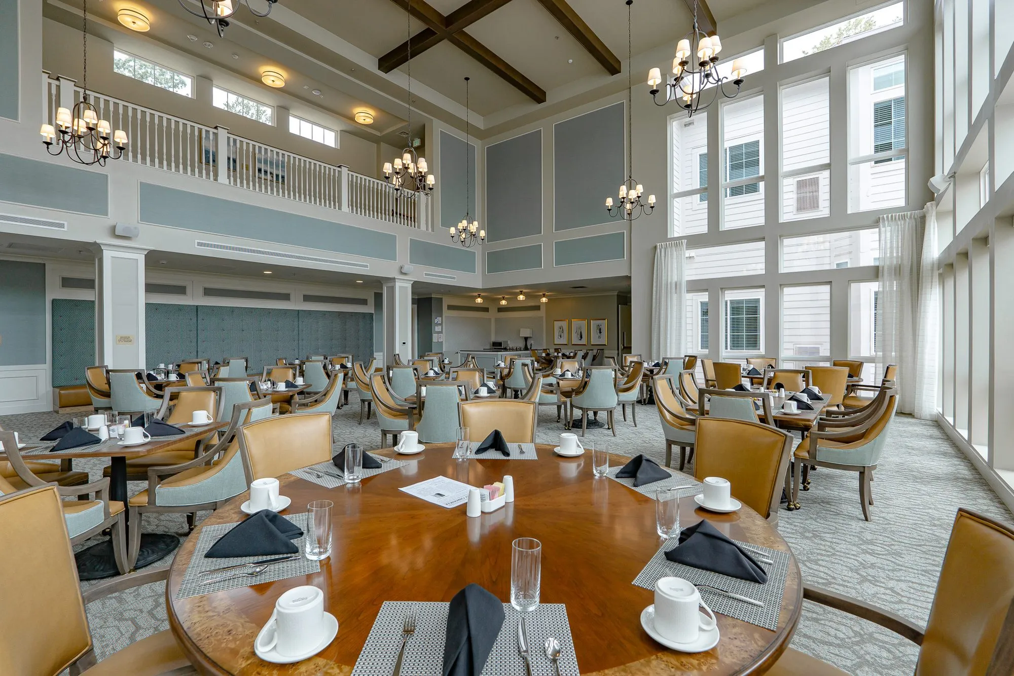 Senior living dining room with a round walnut table set with black napkins and white cups, the beams and balcony beyond