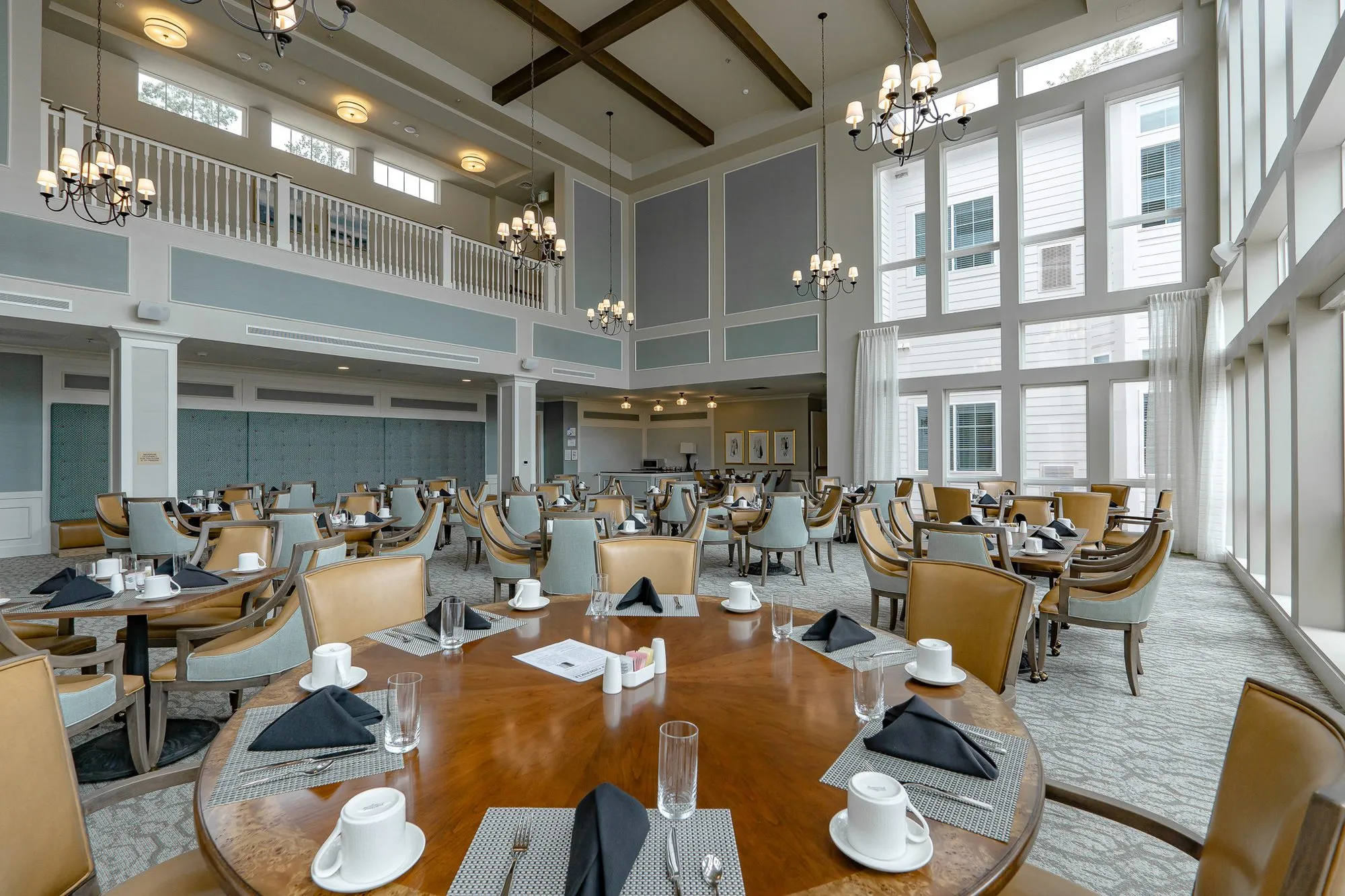 Senior living dining room seen over a set round table, tall windows at right and the balcony with chandeliers at left
