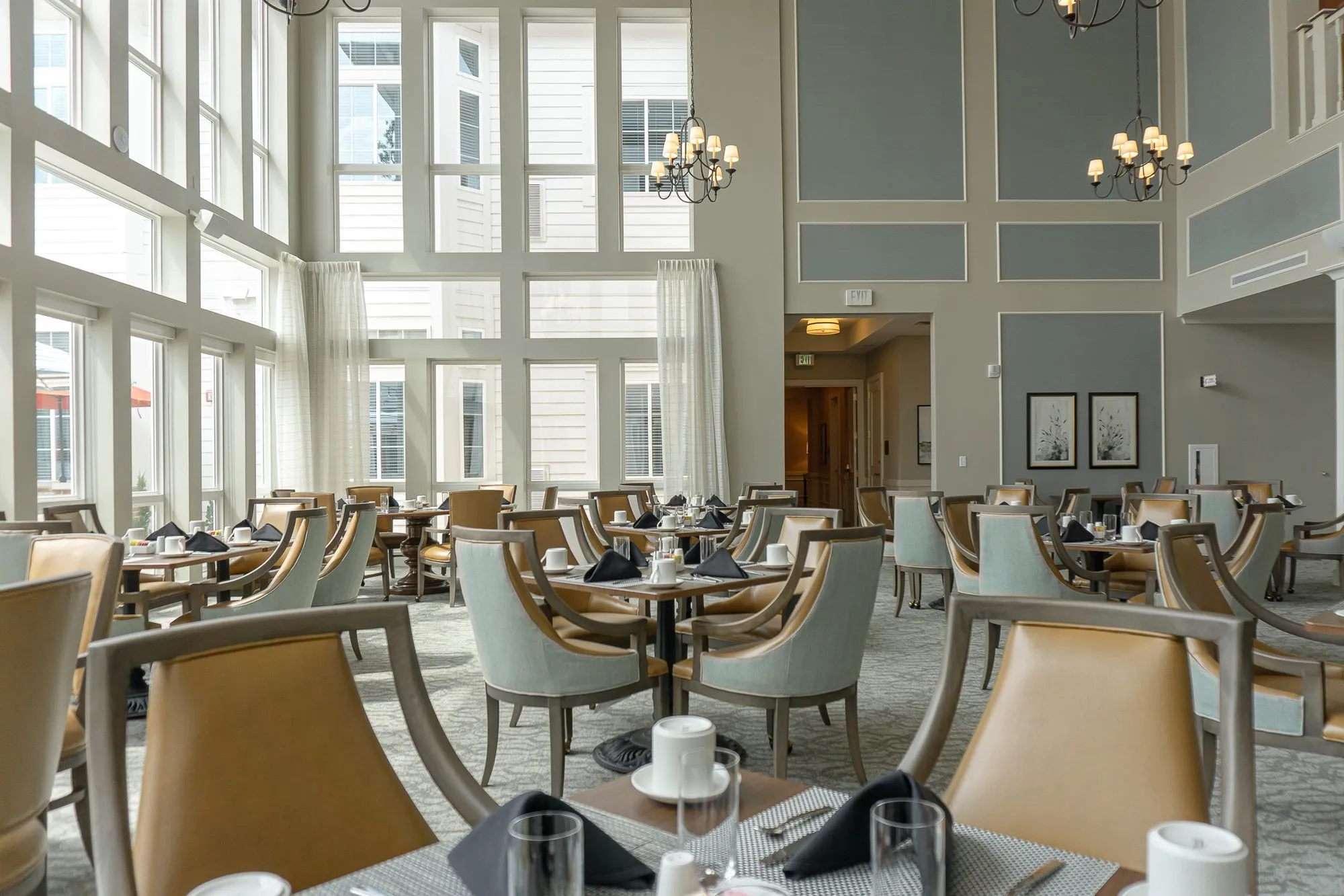 Senior living dining room with a two storey window wall behind sage and camel chairs at set tables