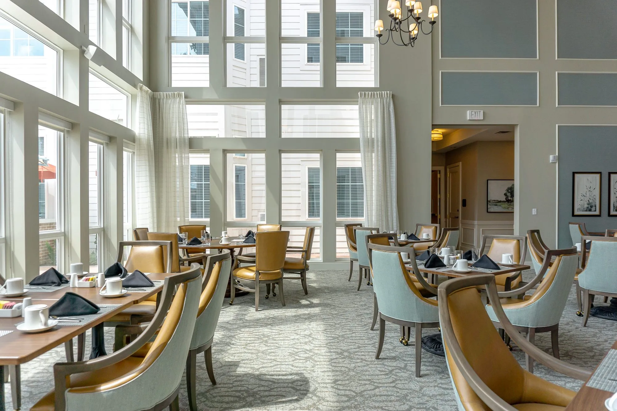 Senior living dining room with a long table set beside two storey windows with sheer curtains, a candle chandelier above