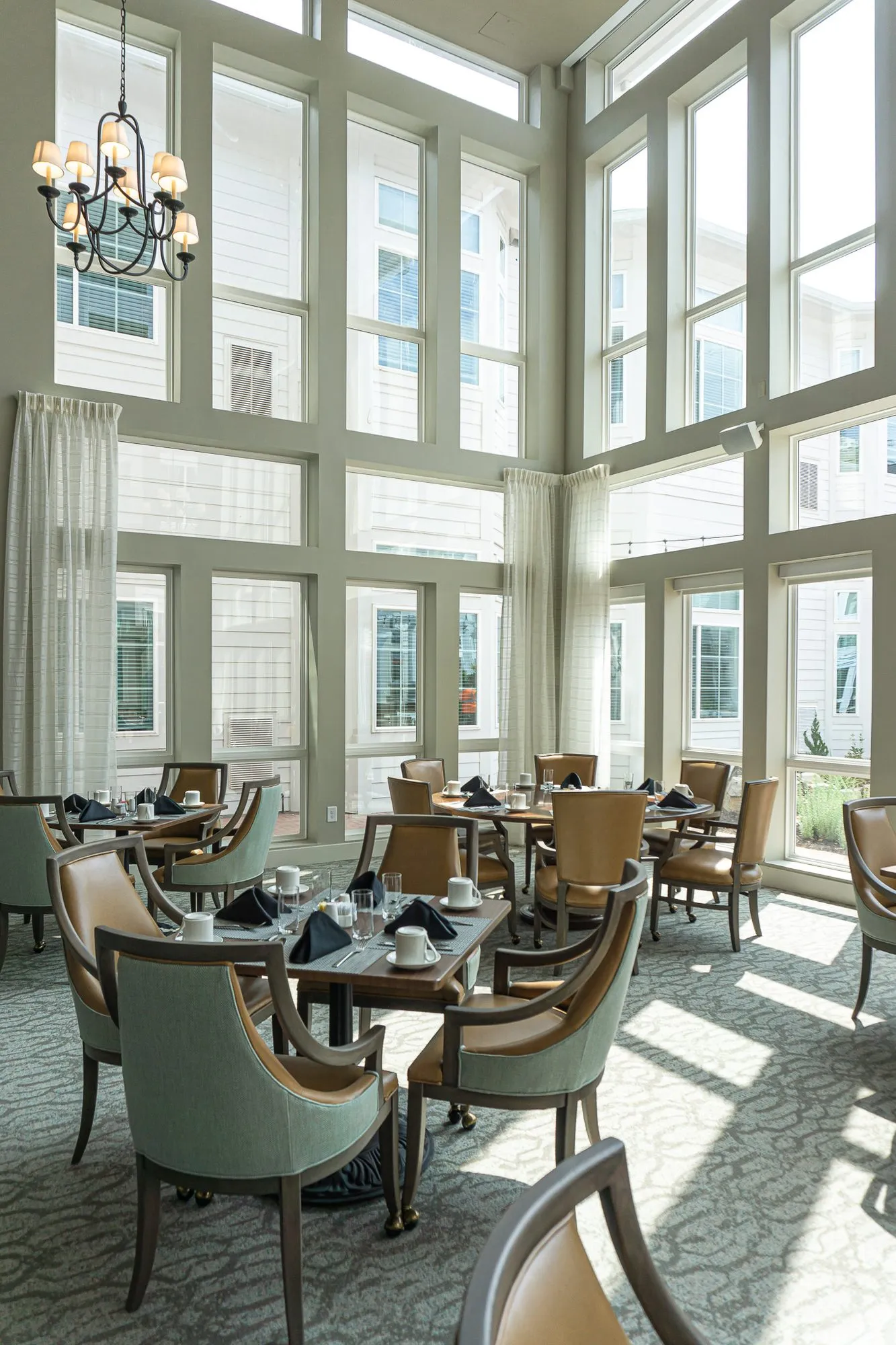Senior living dining room with set tables in a sunlit corner of tall windows under a candle chandelier