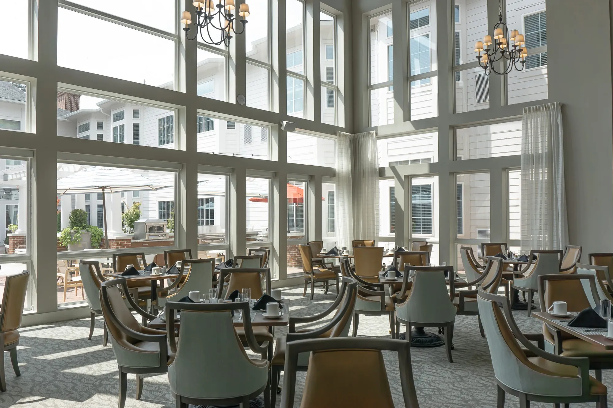 Senior living dining room with sunlit tables before a two storey window wall looking onto courtyard umbrellas