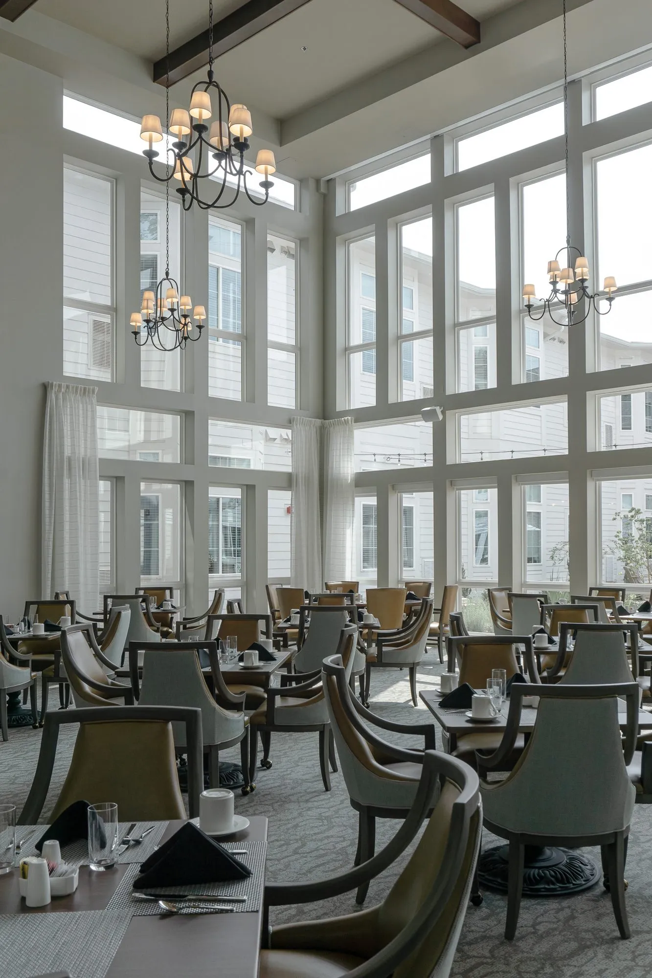 Senior living dining room with tall windows and candle chandeliers above rows of set dining tables