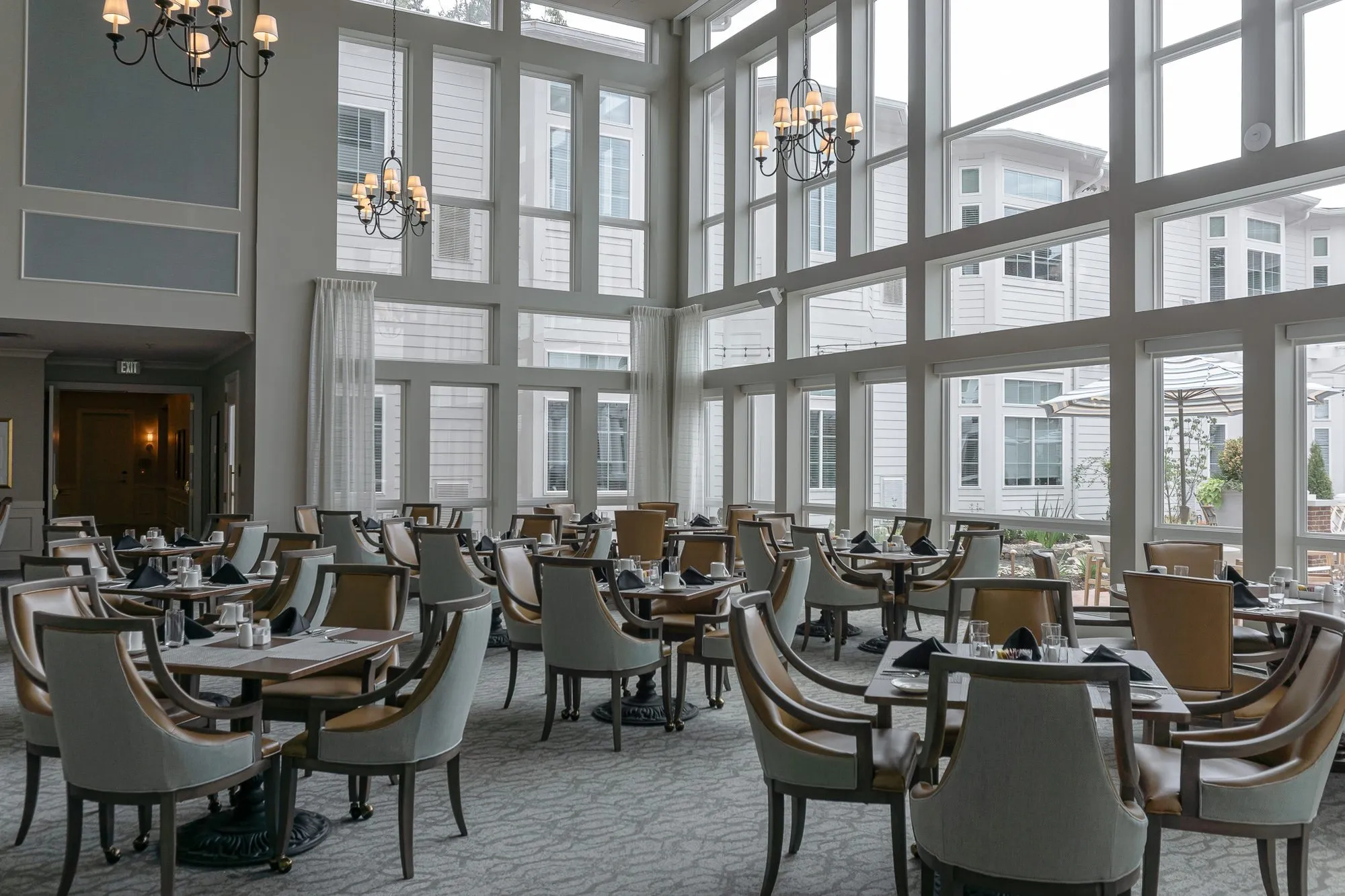 Senior living dining room with rows of tables, sage and camel chairs below a two storey window wall and a wood doorway