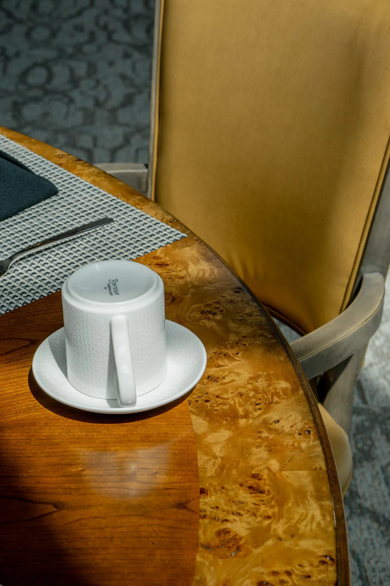 Senior living interior detail: white cup on a saucer at the edge of a burl wood table beside a camel chair