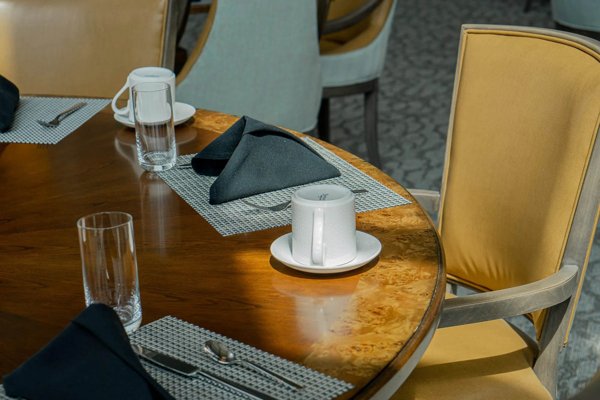 Senior living interior detail: burl wood table set with black folded napkins, cups and glasses beside a camel chair