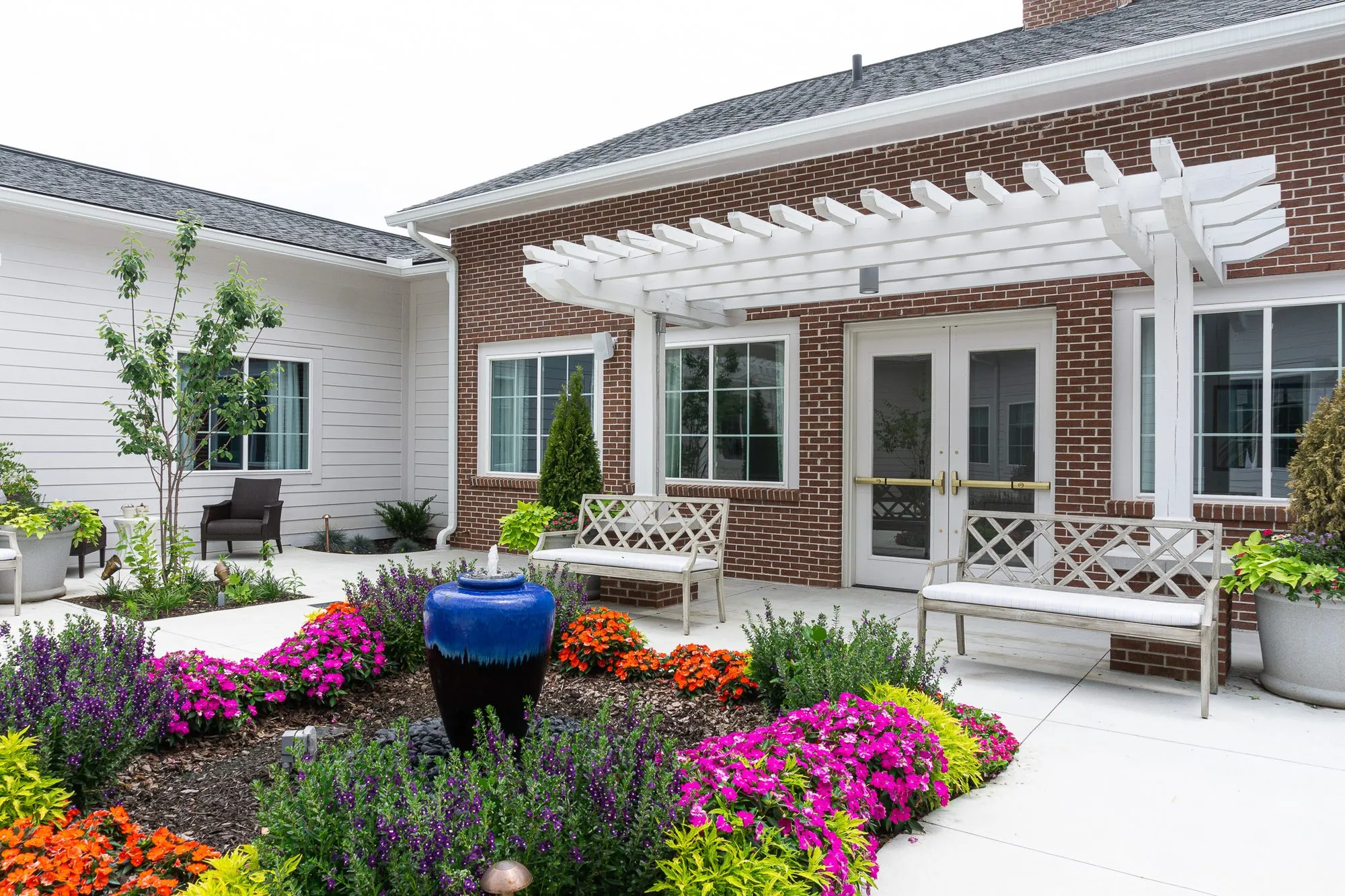 Senior living courtyard garden with a blue glazed fountain urn among pink and orange flowers, white benches under a pergola