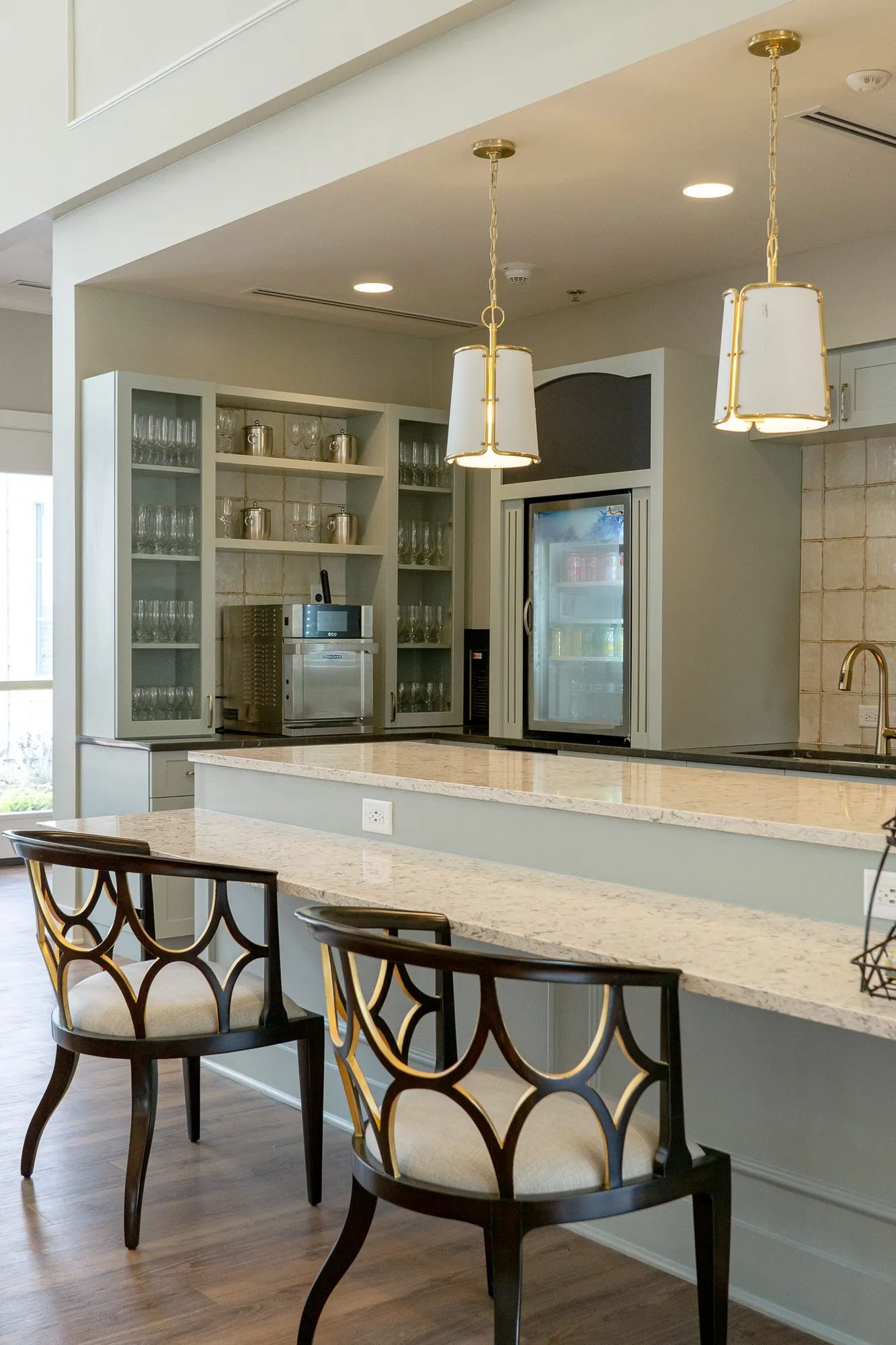 Senior living kitchen with two counter chairs at the stone island under linen pendants, glass cabinets and a cooler beyond