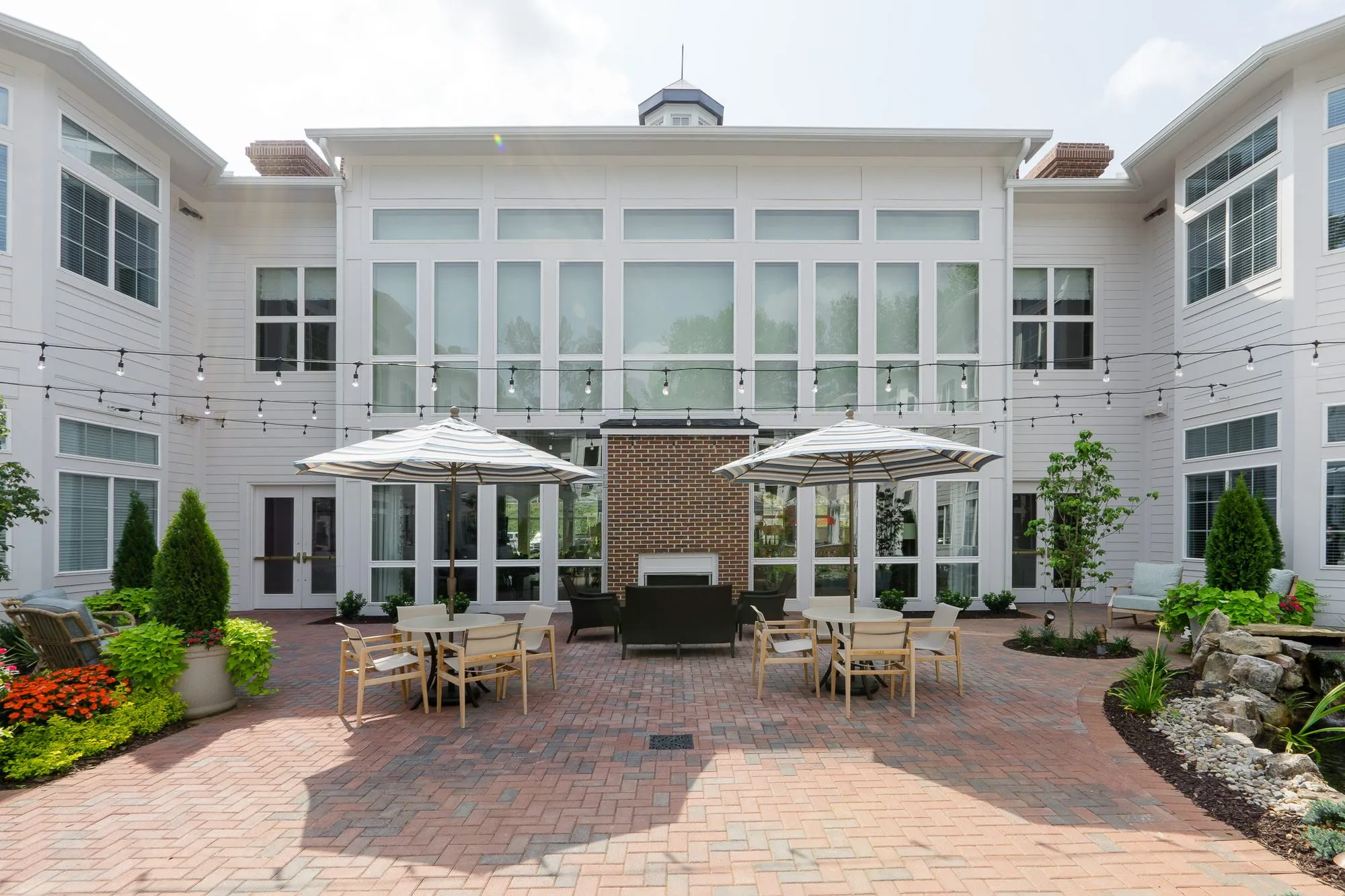 Senior living courtyard with umbrella-shaded tables before a brick outdoor fireplace and the glass-fronted central wing