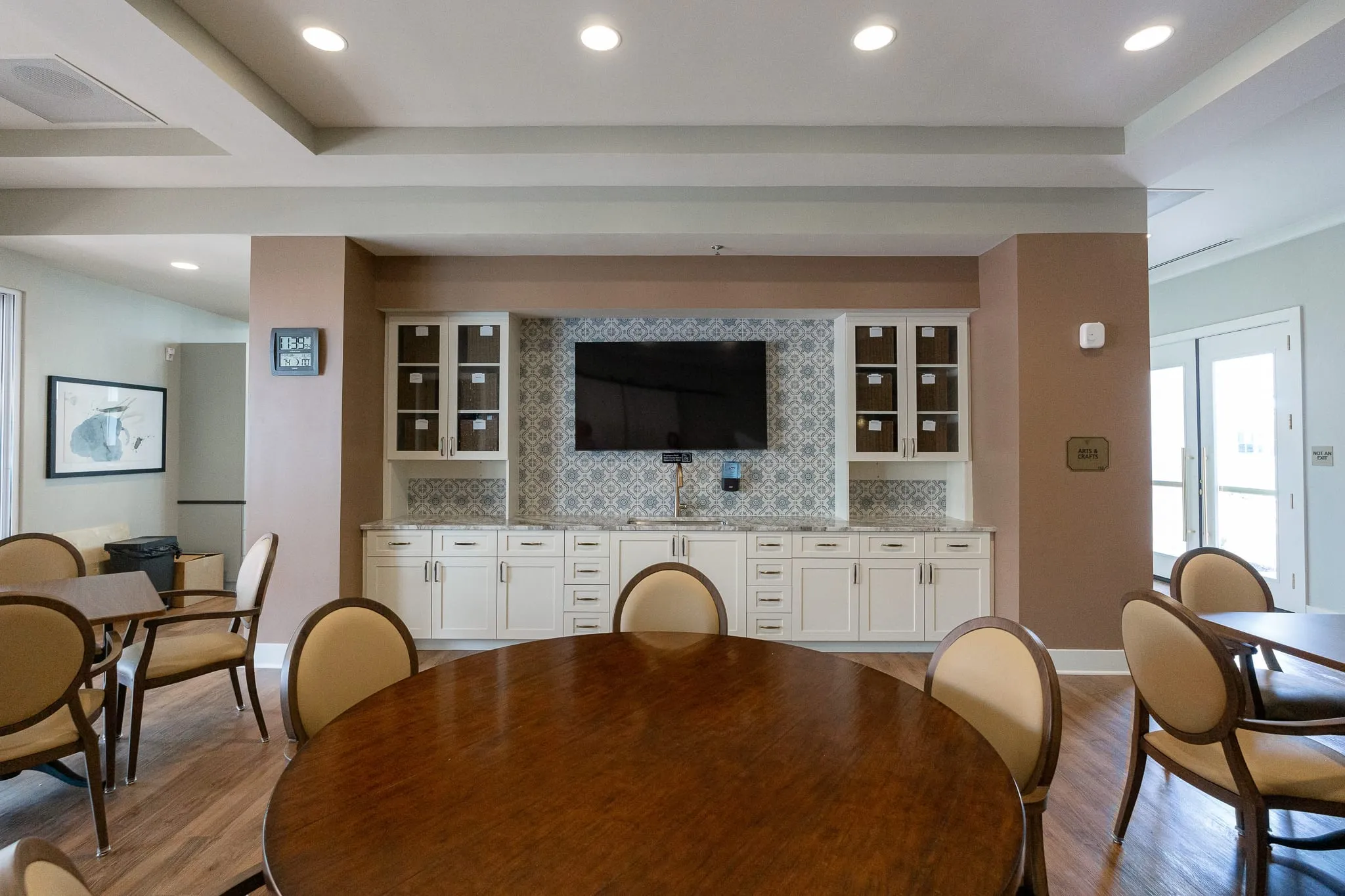 Senior living activity room with cream upholstered chairs at round wood tables facing built-in cabinetry and a TV