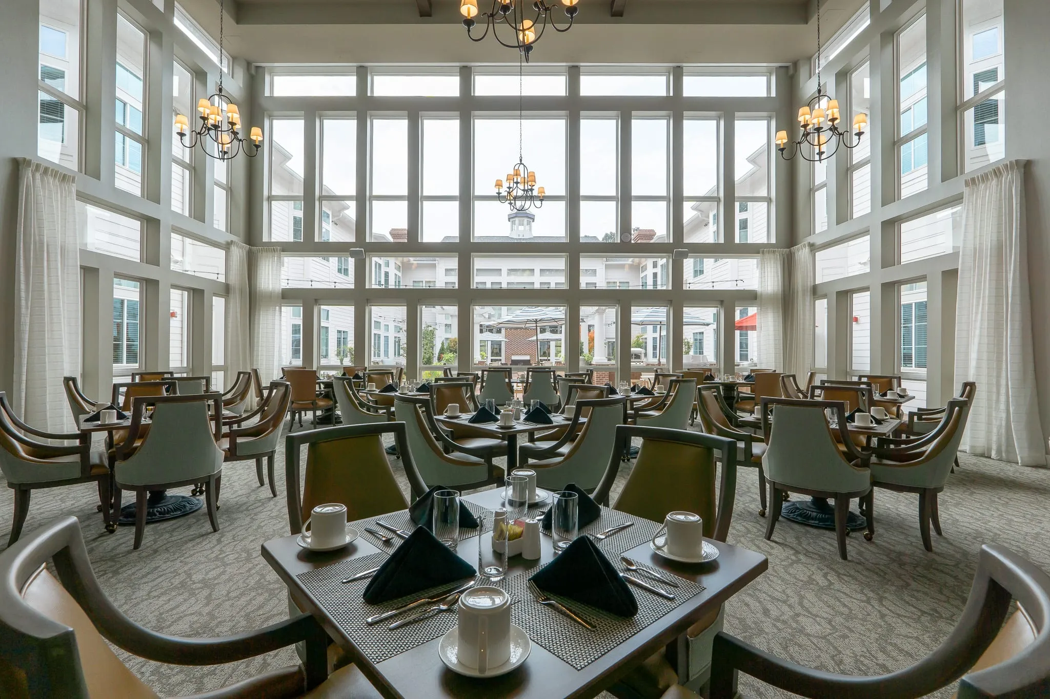 Senior living grand dining room with a two-story window wall and iron chandeliers above set tables, sage and camel chairs