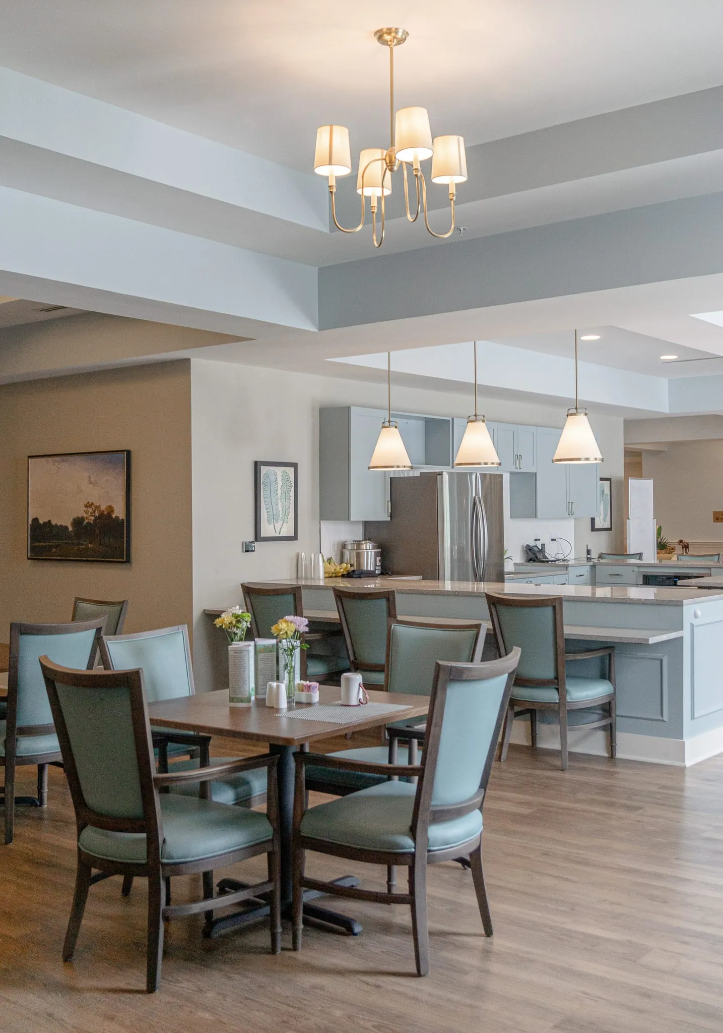 Senior living dining area with a table for four under a chandelier, a kitchen island with stools beyond