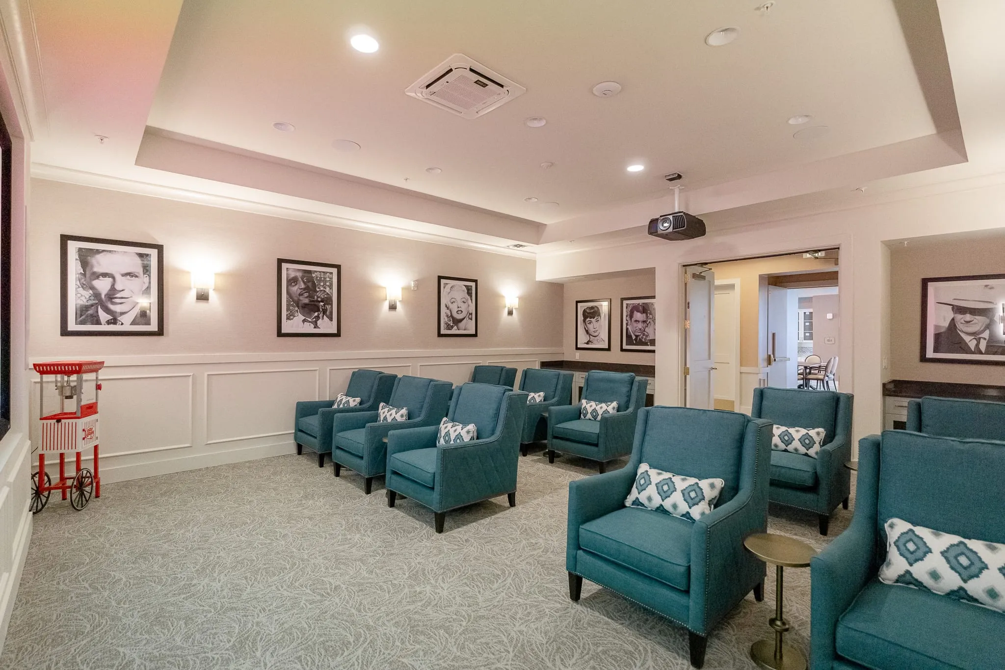 Senior living theater with rows of teal armchairs beneath framed black and white film portraits, a red popcorn cart at left