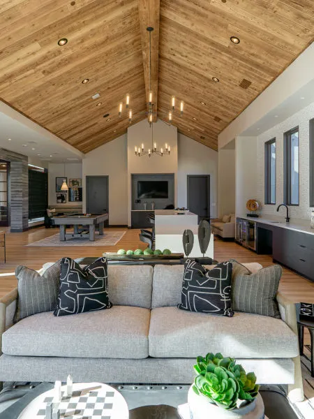 Great room under a vaulted plank ceiling with a linear candle chandelier, a grey sofa in front and a pool table and bar