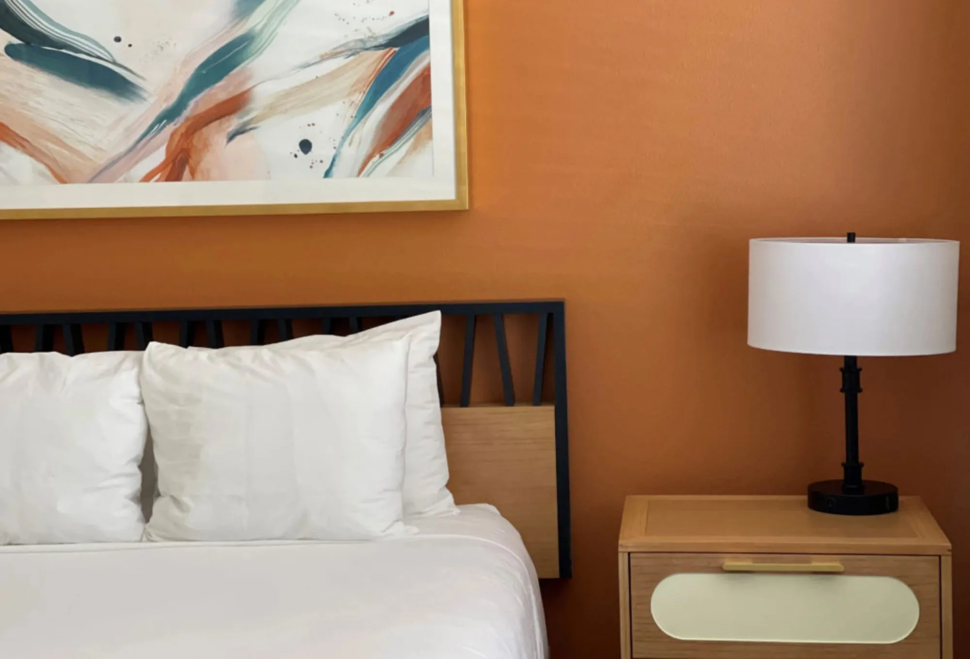 Resort guest room with a black spindle headboard and white bedding against a burnt orange wall, a teal and orange abstract